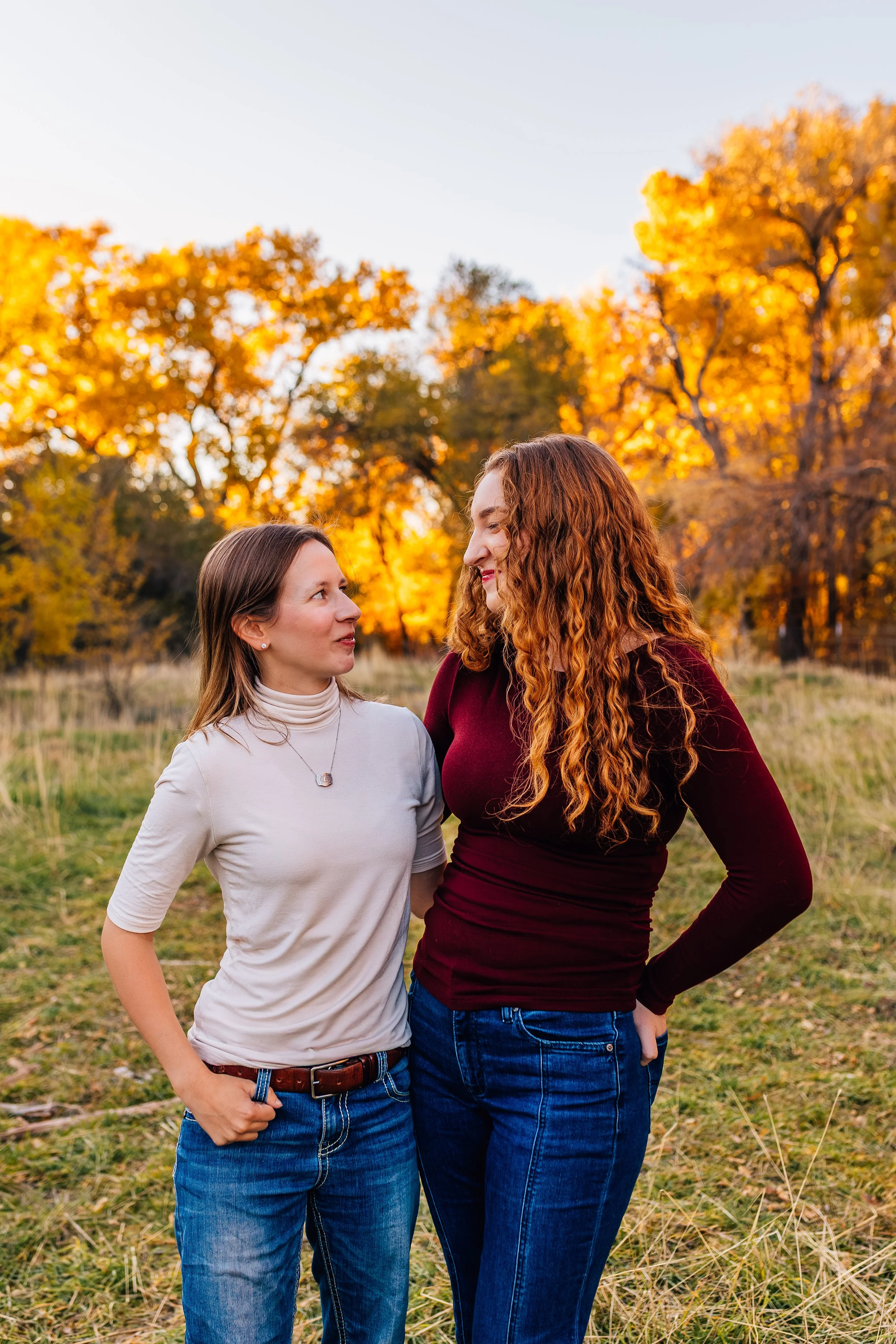 Two women standing close together in an outdoor field during autumn, with trees that have orange and yellow leaves in the background, sharing a moment of closeness.