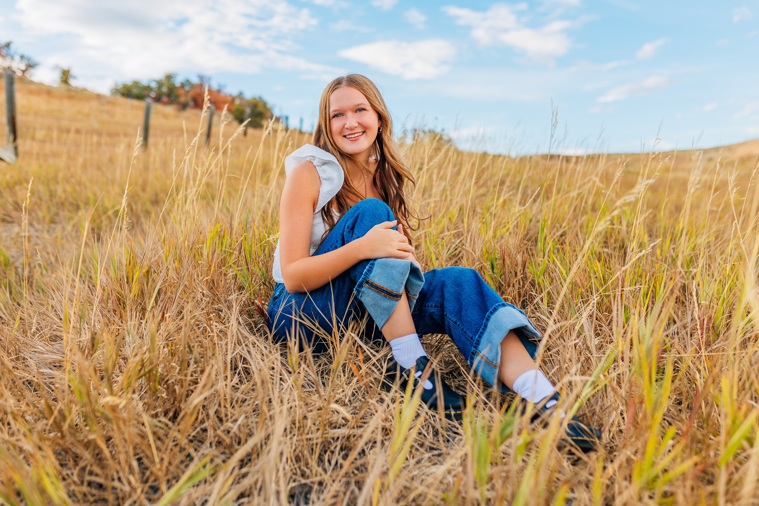 A girl sitting in a field of tall, dry grass, smiling at the camera, with a fence and a blue sky with clouds in the background.