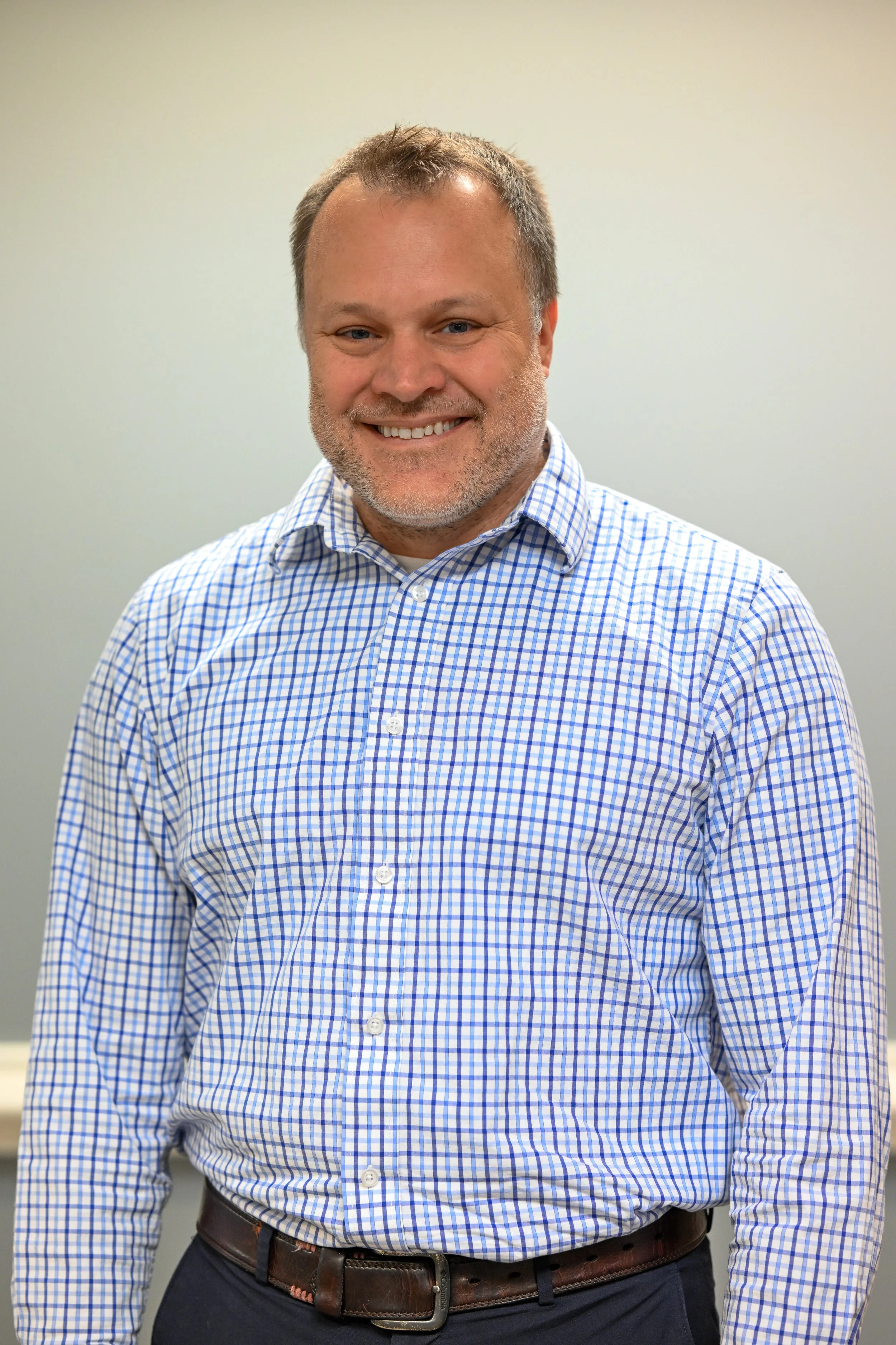 Man smiling, wearing a blue and white checkered shirt, standing against a plain light-colored wall.