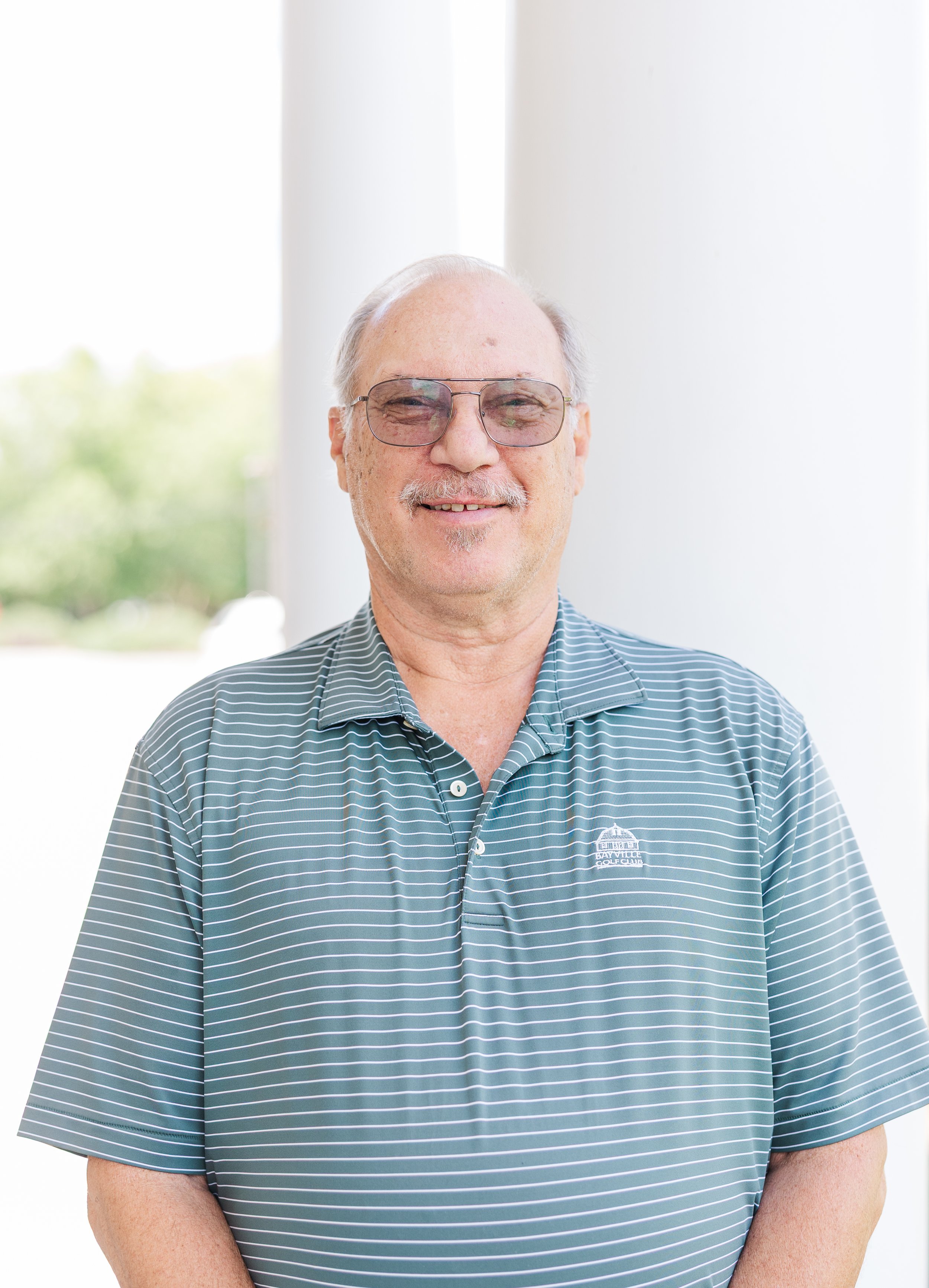 A smiling elderly man with gray hair and glasses, wearing a striped golf shirt, standing outdoors with white columns in the background.