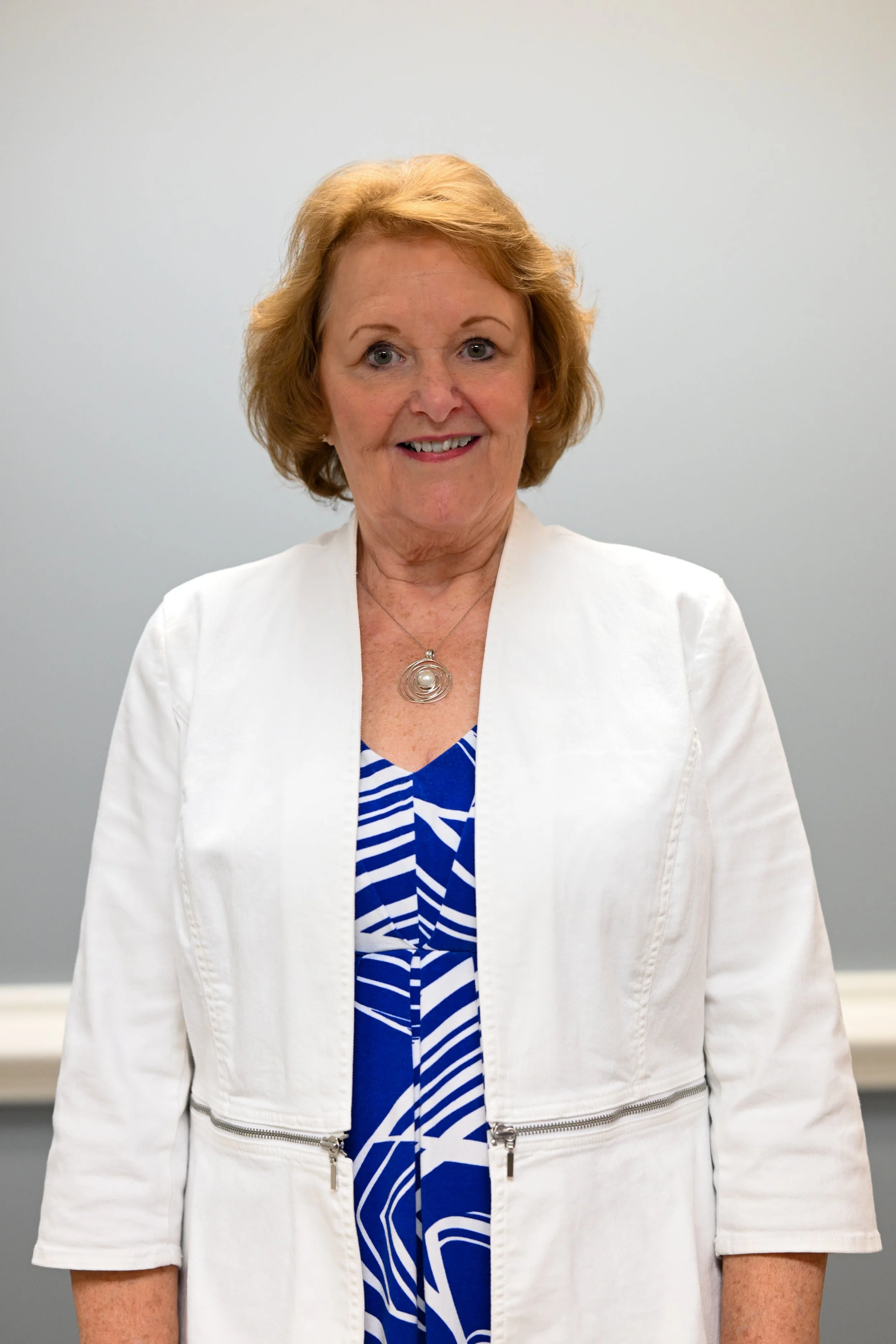 A smiling middle-aged woman with short red hair, wearing a white jacket and a blue and white patterned top, standing against a plain gray background.