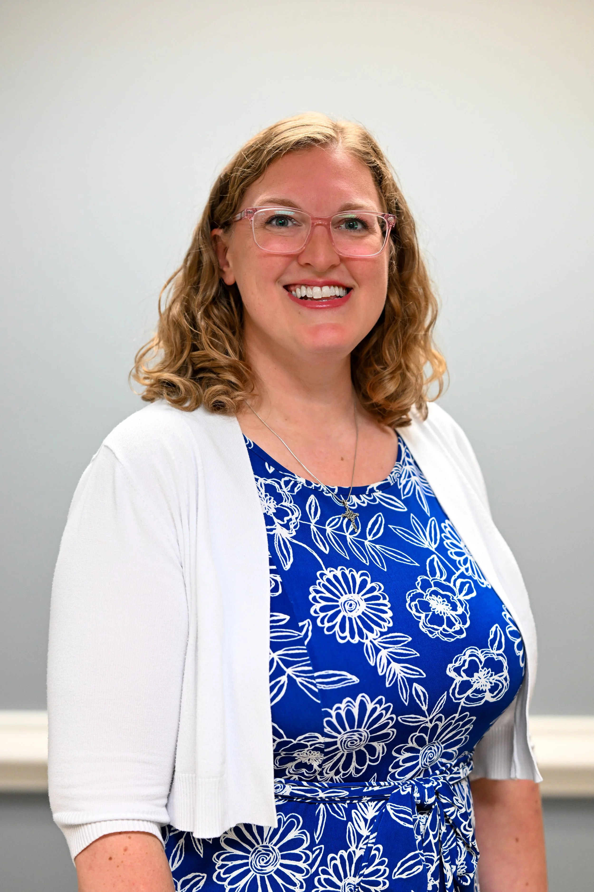 A woman with curly red hair and glasses smiling, wearing a white cardigan over a blue floral dress with a matching belt, in front of a plain gray background.