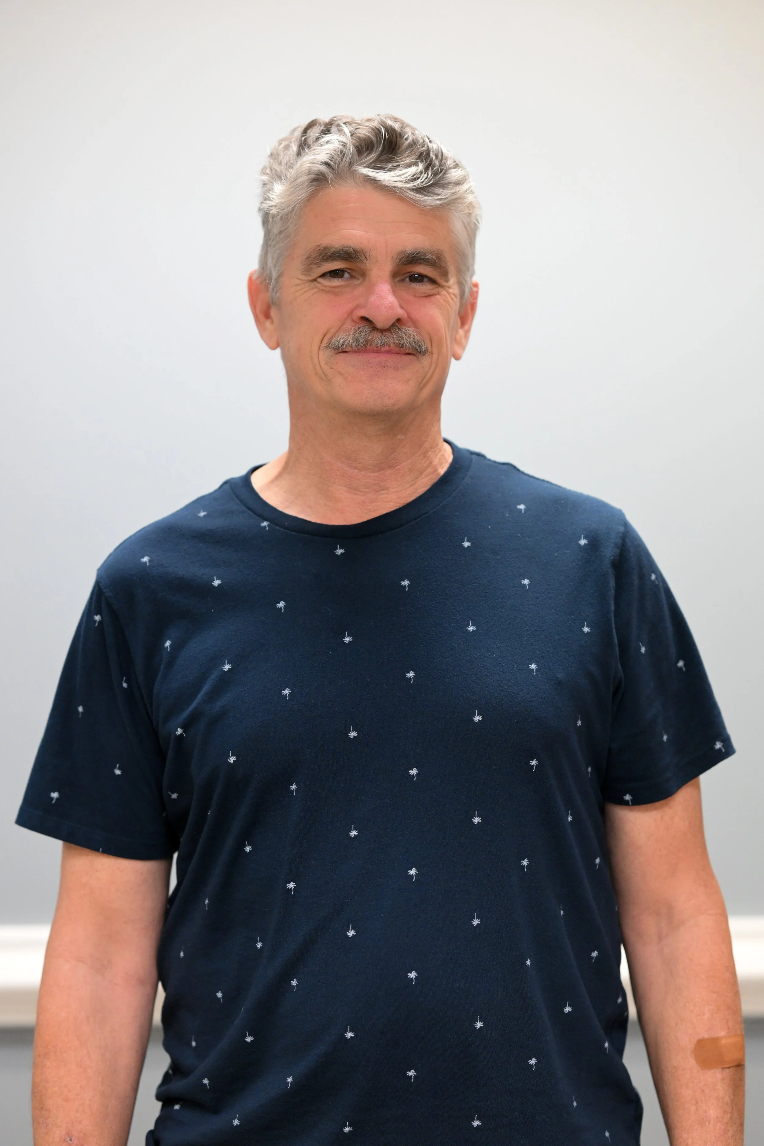 A middle-aged man with gray hair and a mustache, smiling, wearing a navy blue T-shirt with small white palm print, standing against a plain light gray background.