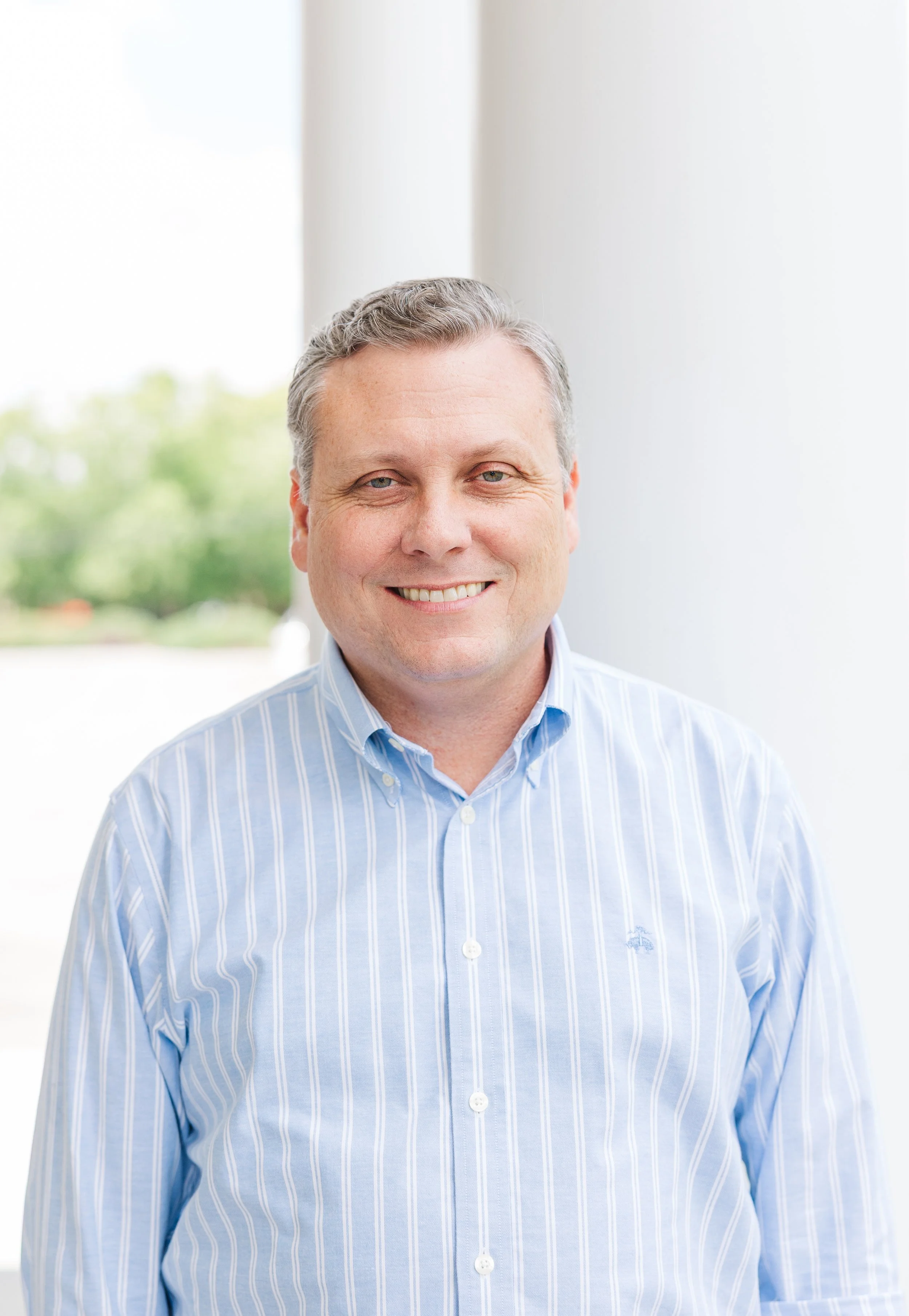 Portrait of a middle-aged man with gray hair, wearing a blue and white striped button-up shirt, smiling at the camera, with a blurred outdoor background.