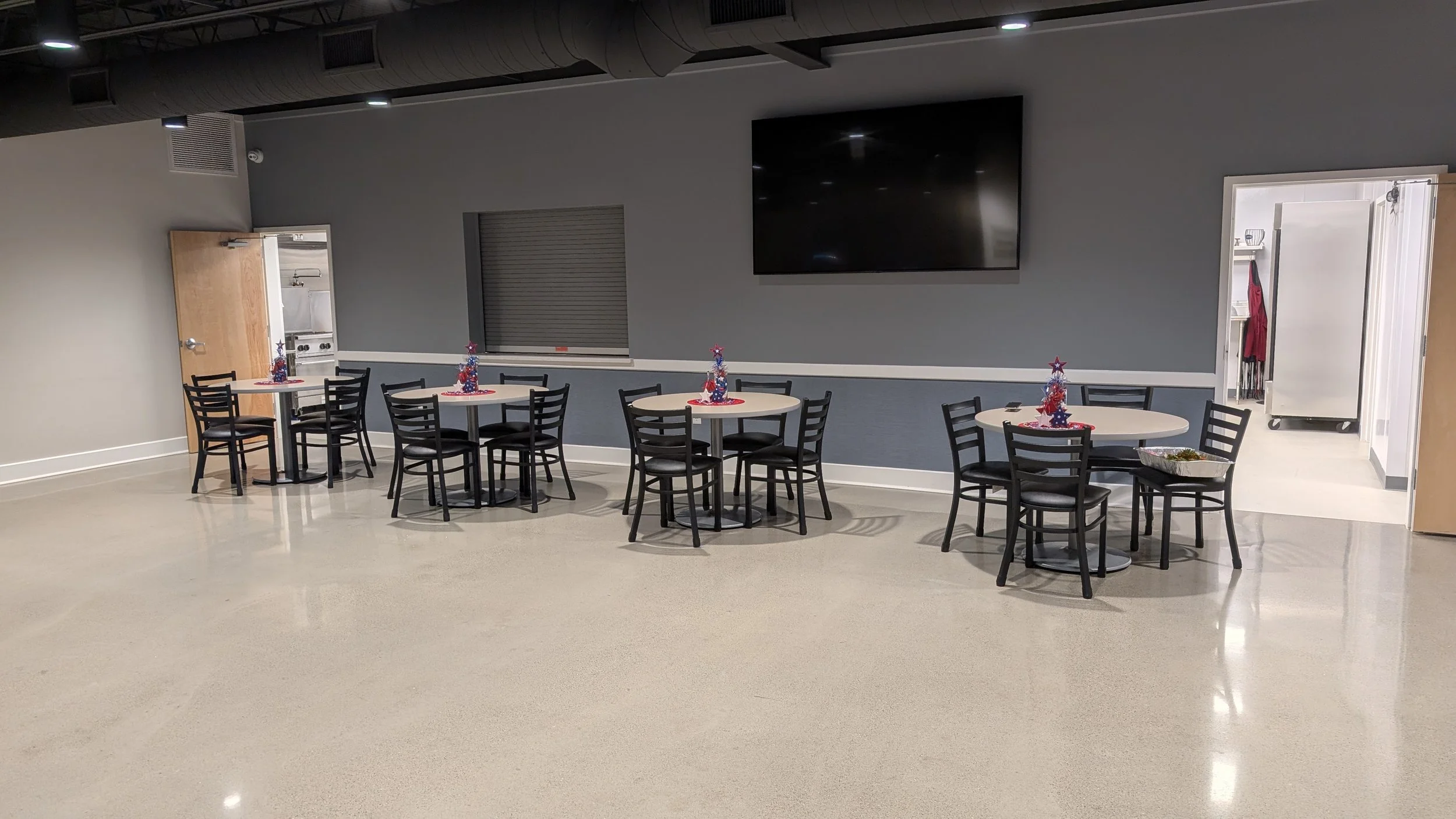 Empty banquet room with four round tables decorated with Christmas-themed centerpieces, black chairs, a large flat-screen TV on the wall, and a doorway leading to a kitchen area.