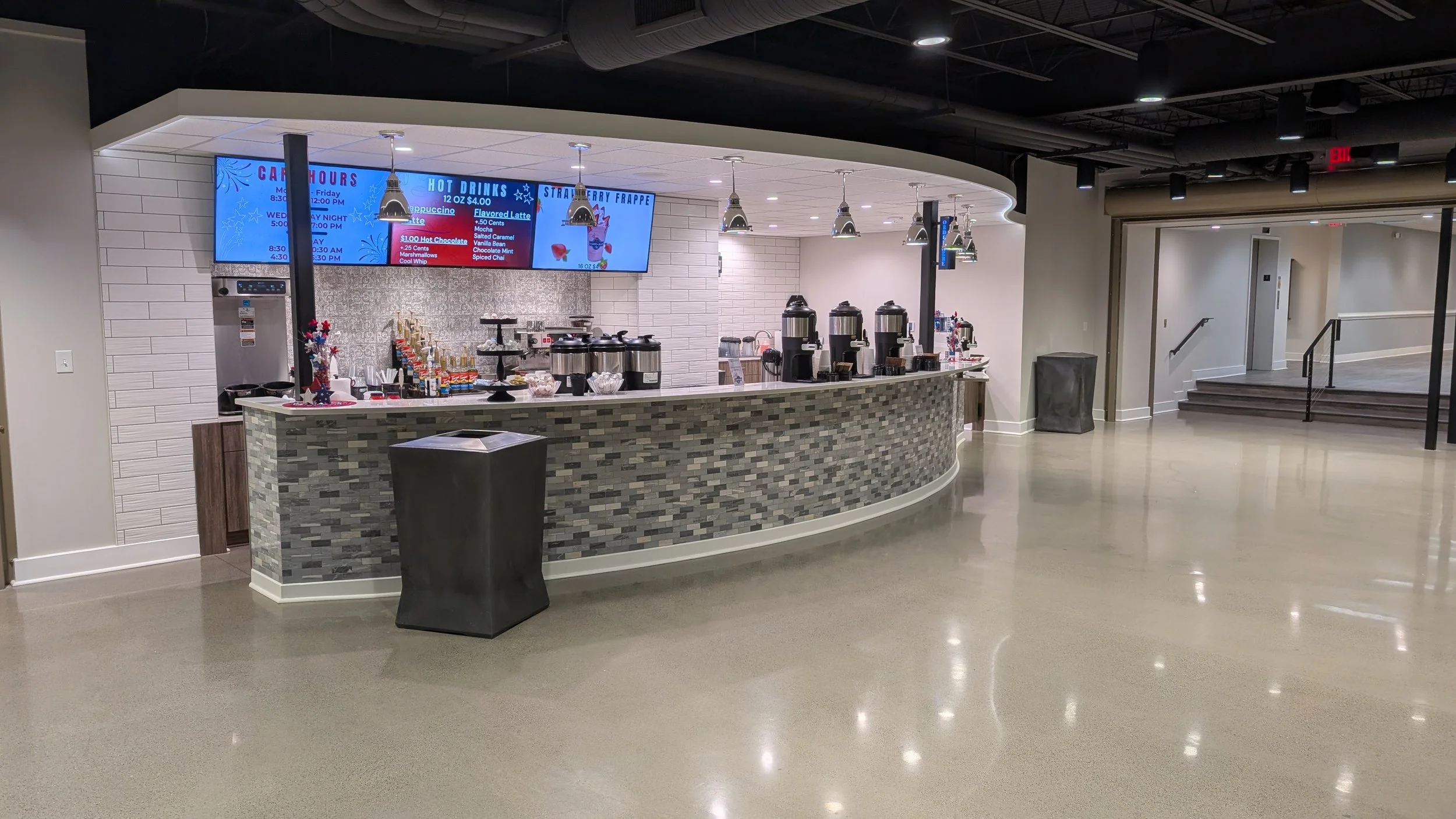 Coffee and beverage station in a modern cafe or lounge area with a curved counter, coffee machines, colorful candy and snack containers, and digital menu screens above. The space has a polished floor and a neutral color palette.