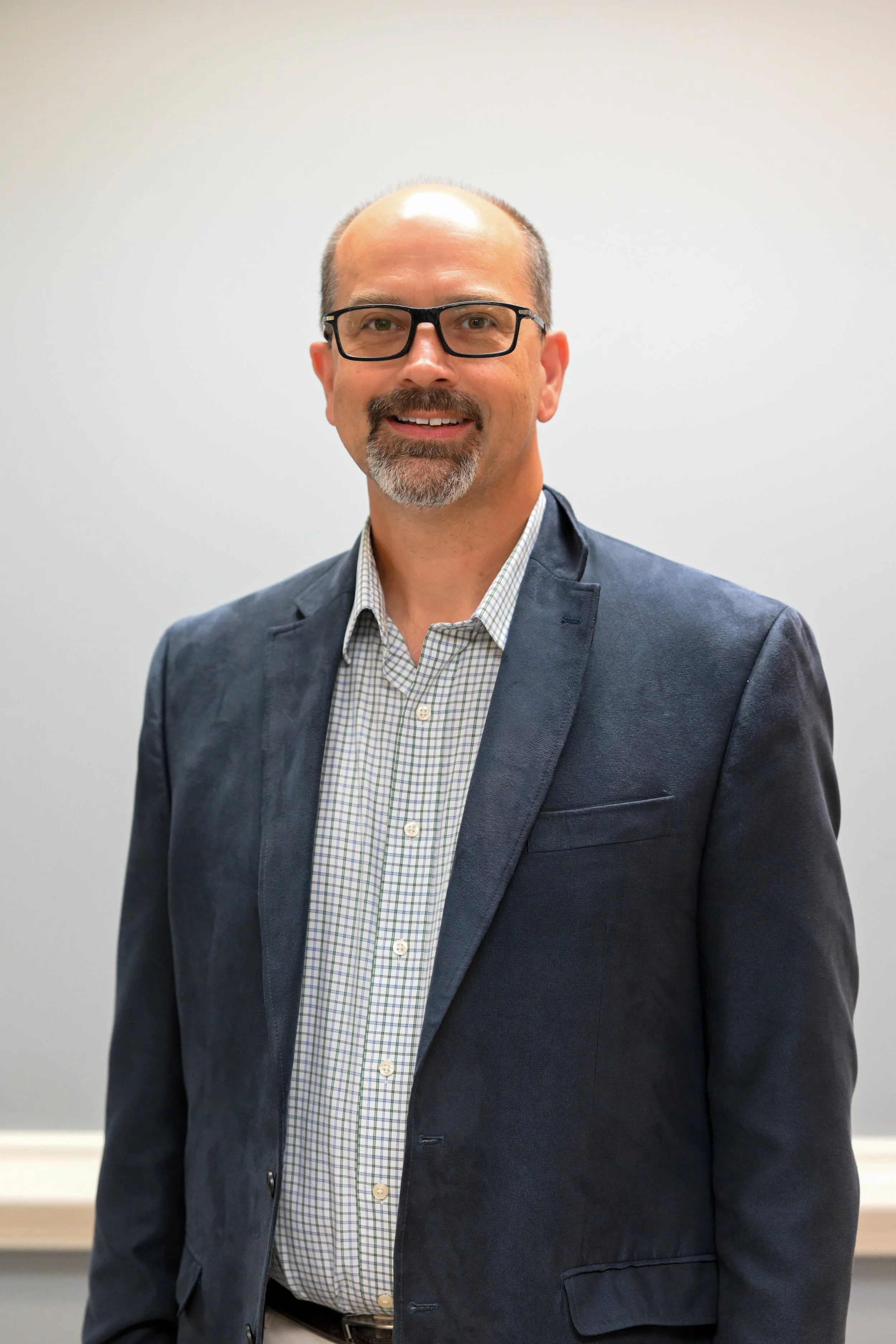 A middle-aged man with glasses, a beard, and a mustache, dressed in a blazer and checked shirt, standing in front of a plain gray wall and smiling.