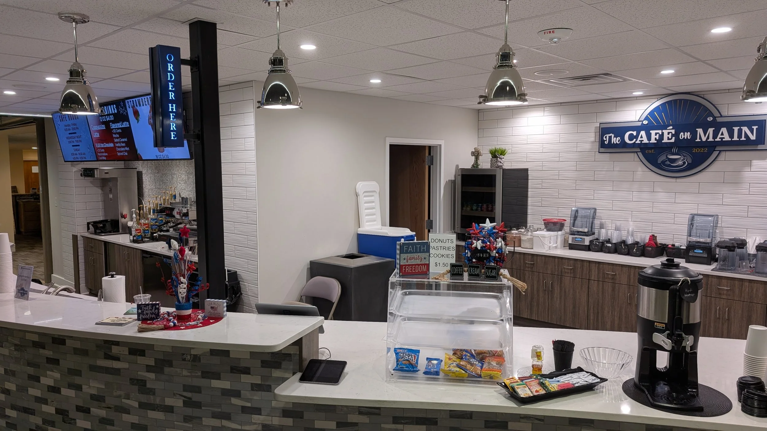 Counter at a cafe with a coffee machine, snack display, and signs about donuts, pastries, and cookies, with a wall sign reading 'The Café on Main' in the background.