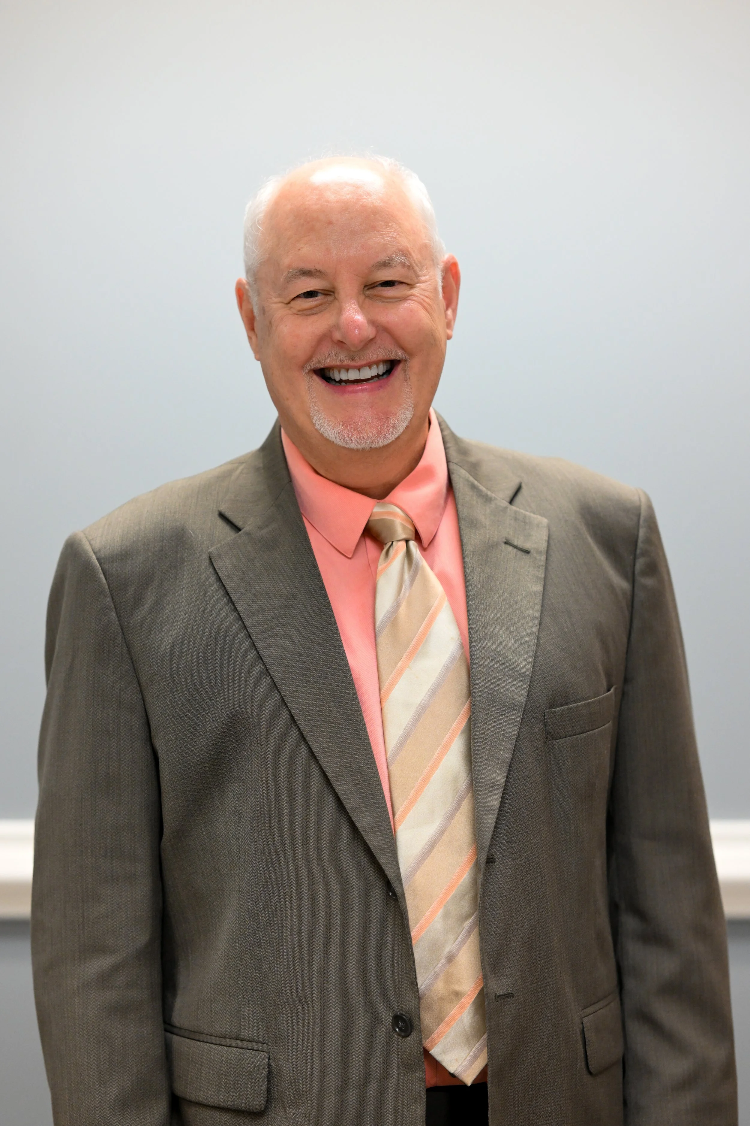 A smiling man with a beard and mustache wearing a gray suit, pink shirt, and a striped tie, standing against a light gray background.