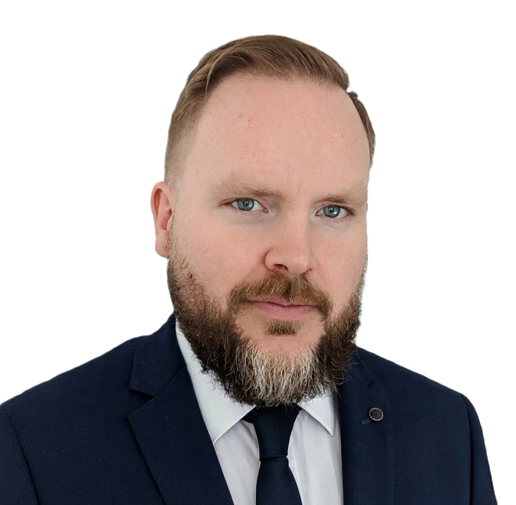 A man with a beard and short blond hair wearing a dark suit, white shirt, and dark tie.