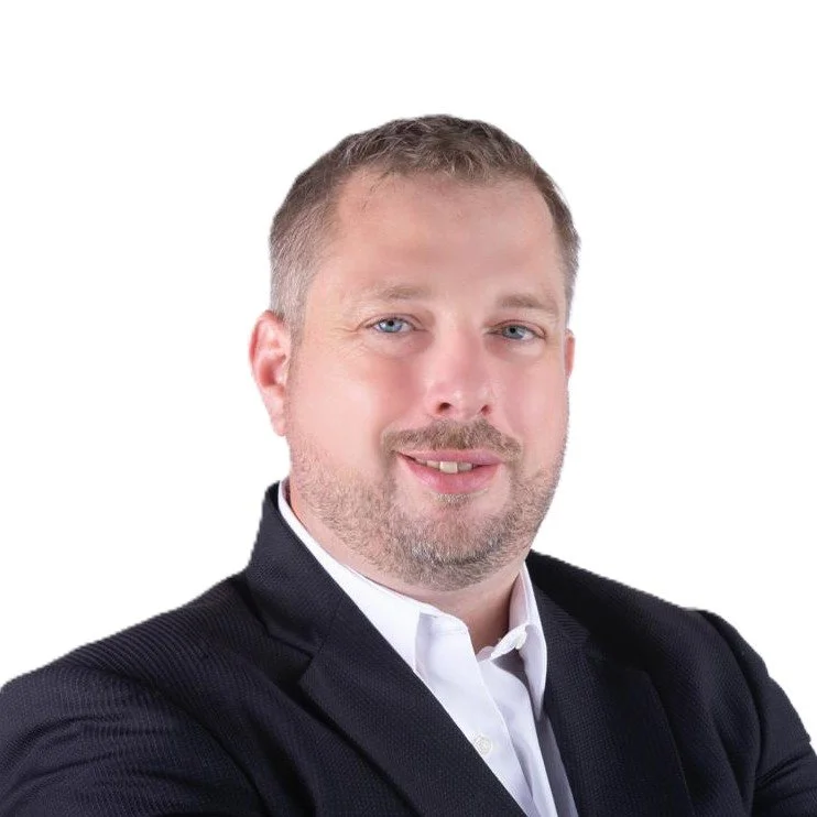 Portrait of a man with light skin, short light brown hair, and blue eyes, wearing a dark blazer and white shirt against a white background.