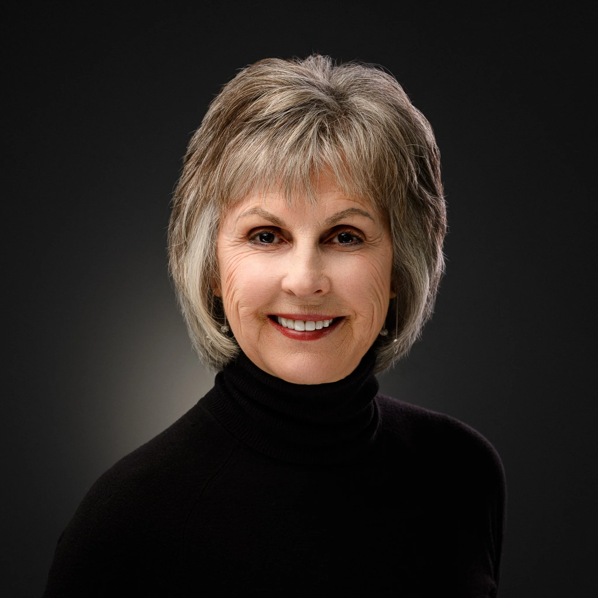 A smiling woman with short blonde hair, wearing a dark blue shirt and earrings, against a gray background.