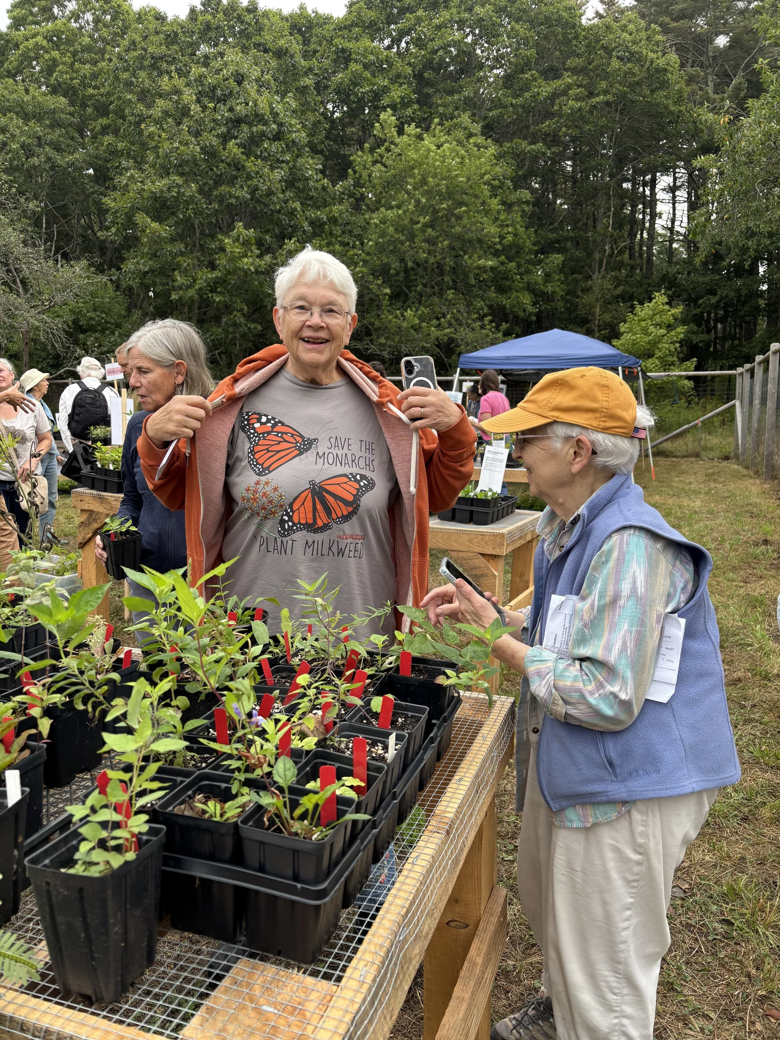 Two people at an outdoor native plant sale with multiple small plants in black containers on a wooden table, surrounded by trees and other visitors.