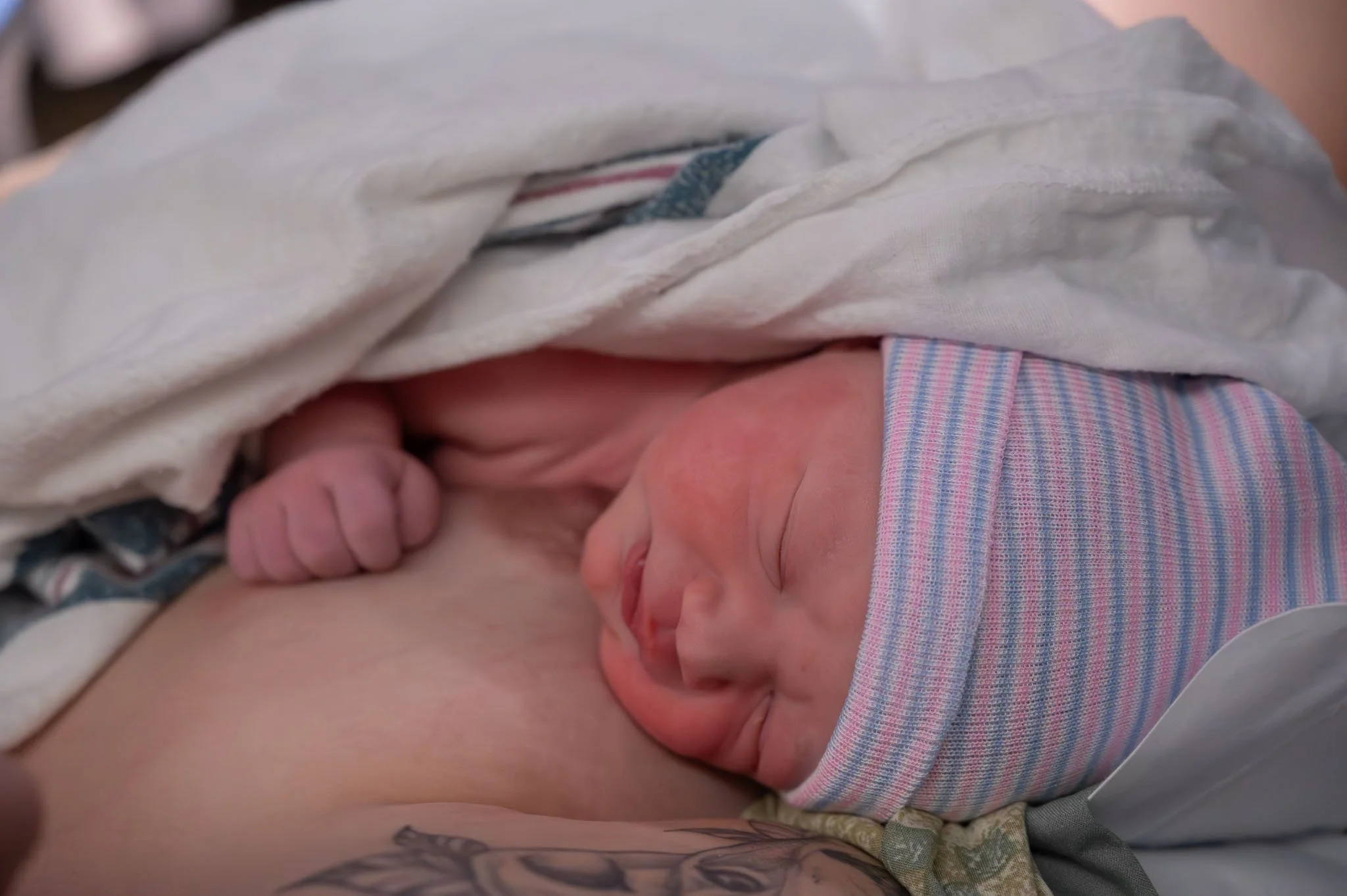 Close-up of a newborn baby wearing a striped hat, lying on an adult's chest, wrapped in a blanket.