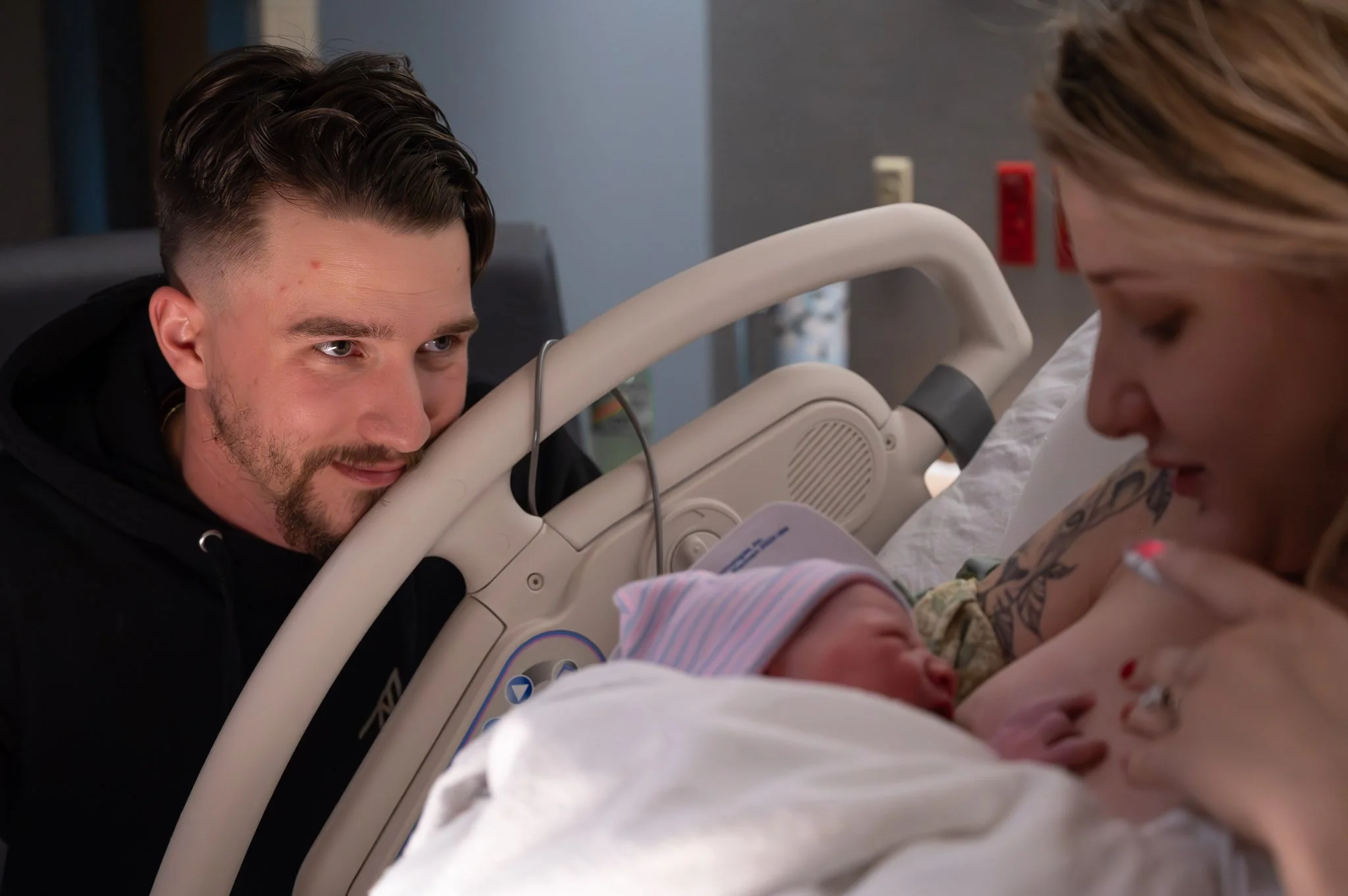 A woman holding a newborn baby in a hospital bed, with a man leaning over the bed, looking at the baby.