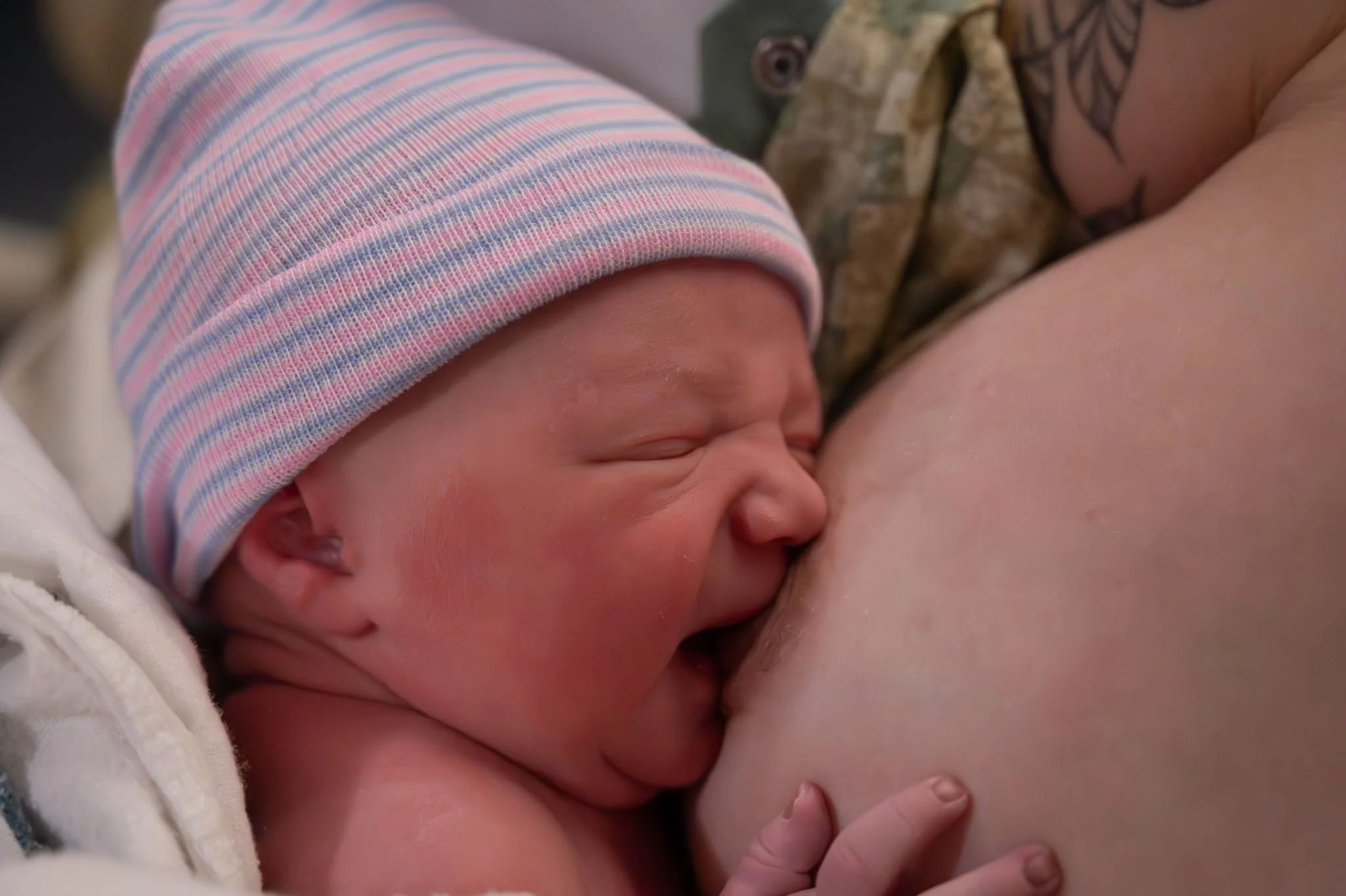 Newborn baby nursing, wearing a pink and blue striped hat, being held by a woman with visible tattoos.