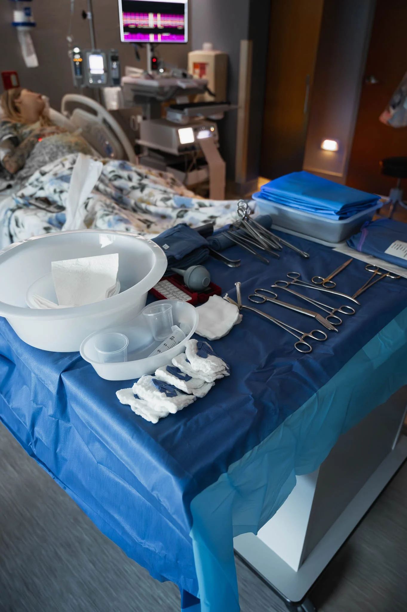 Medical instruments, supplies, and sterile drapes arranged on a blue table cover in a hospital room with a patient in bed.