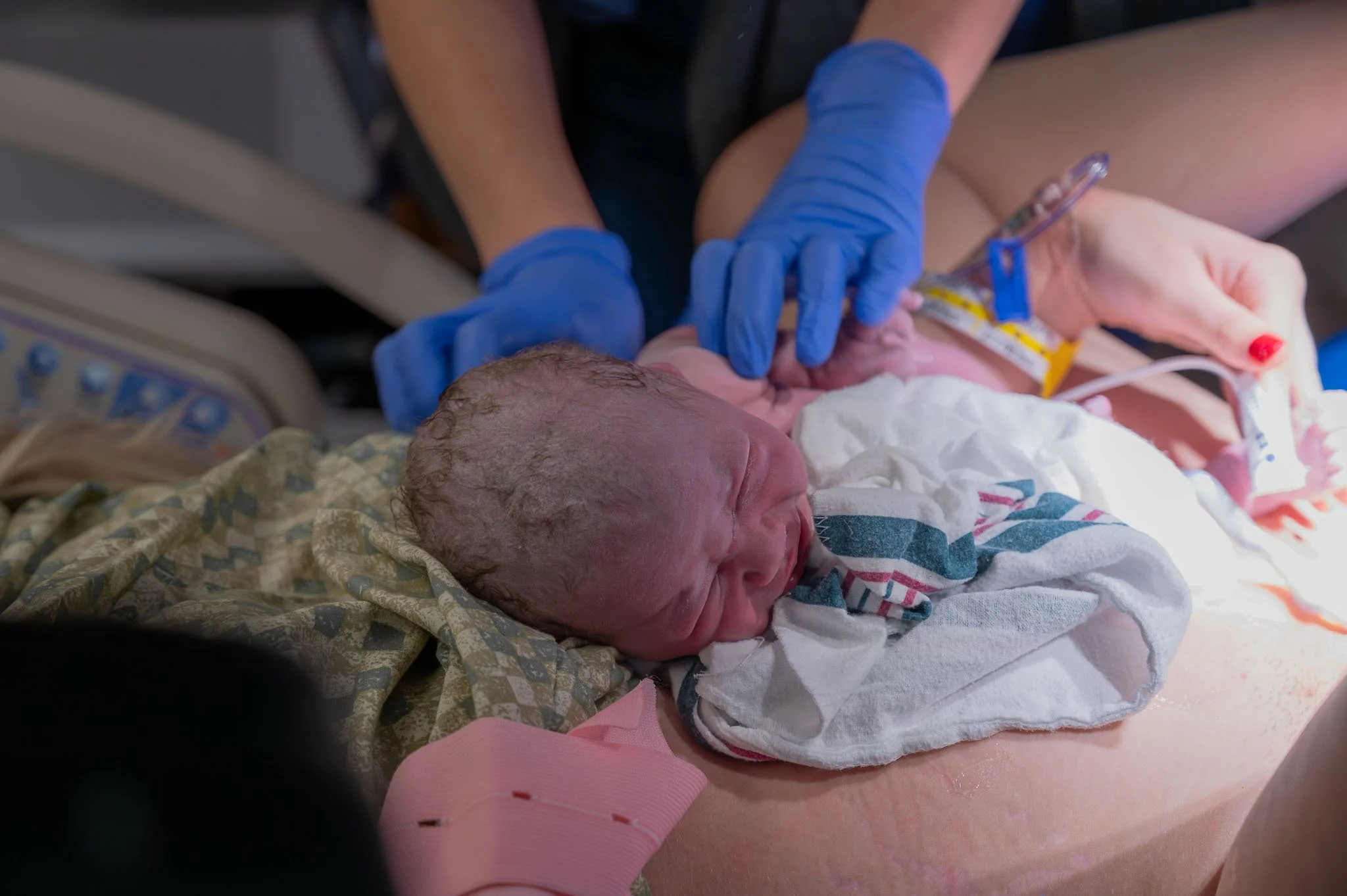 A newborn baby with wet hair lying on a hospital bed, surrounded by medical personnel wearing blue gloves, holding the baby's hand and head during birth.