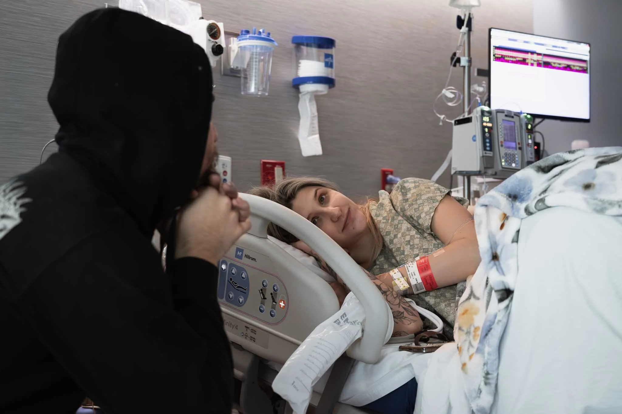 A woman lying in a hospital bed looking at a person sitting beside her, holding her hand, in a hospital room with medical equipment and monitors on the wall.
