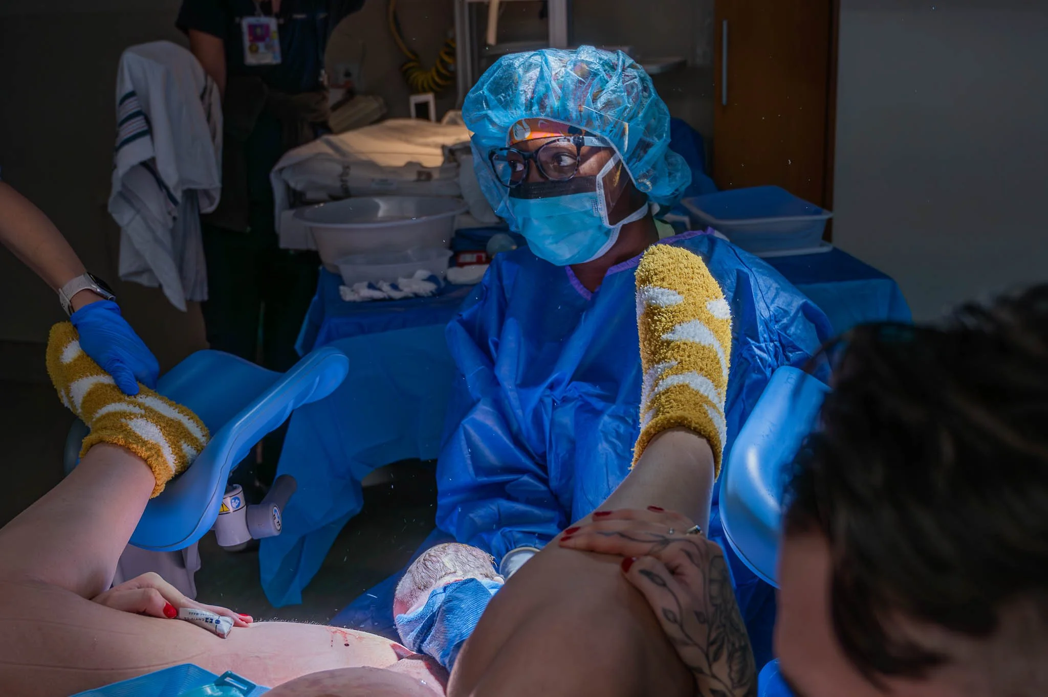 Medical professional in surgical gown, mask, gloves, and protective eyewear assisting during childbirth in hospital setting.