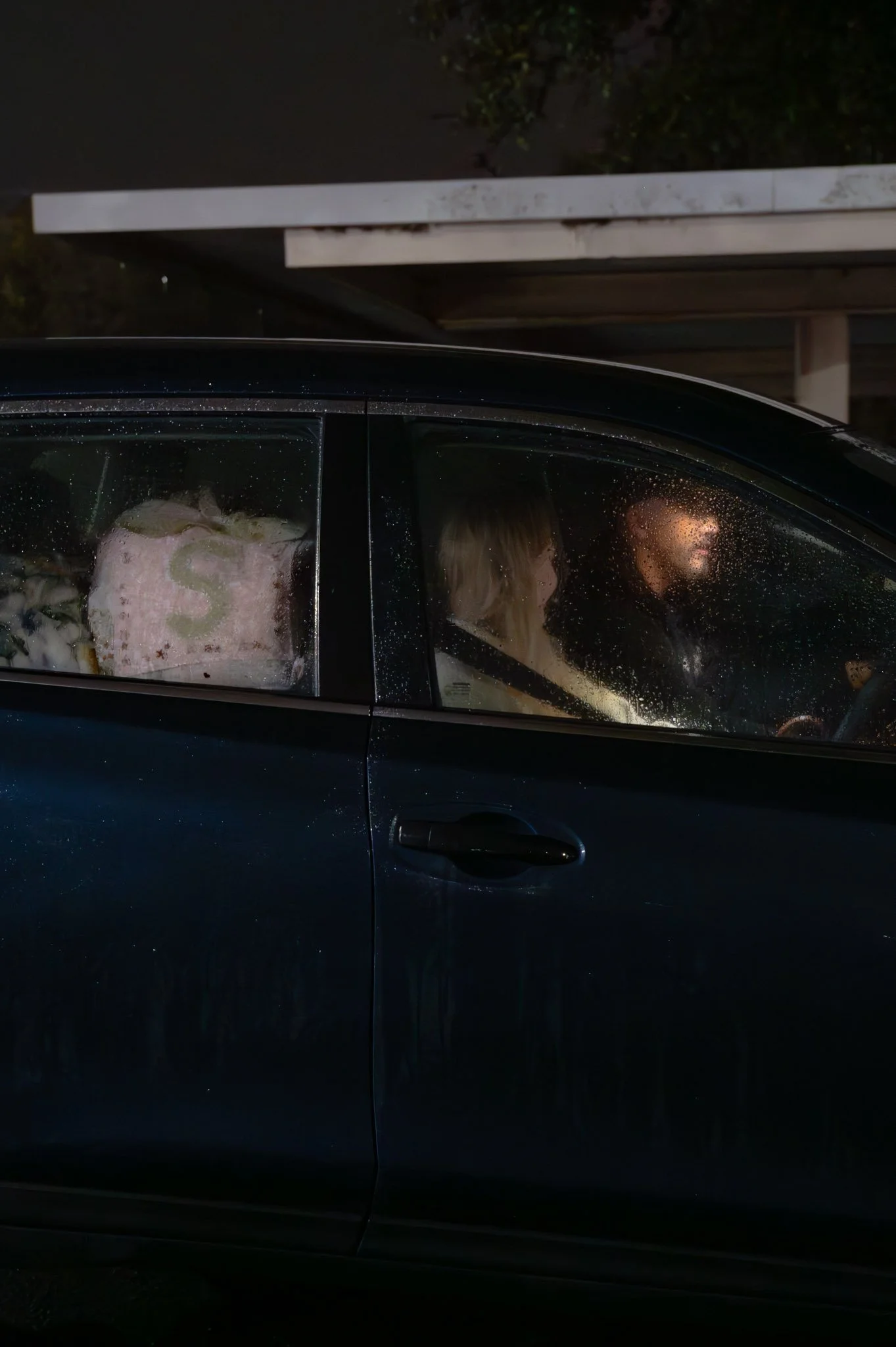A black car parked outdoors at night with rain on the windows. Two people sit inside, a woman with blonde hair and a man, visible through the wet glass.
