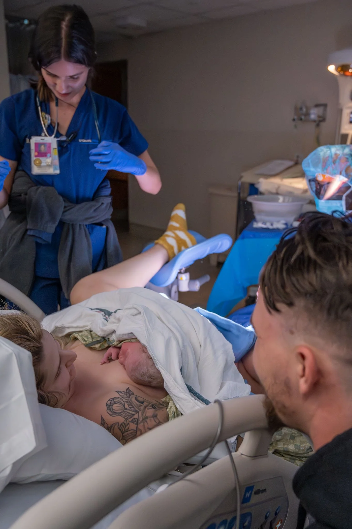 A woman in labor lying in a hospital bed with a partner close by, while a medical professional prepares to assist. The woman has tattoos on her chest, and a newborn baby is lying on her chest. Medical staff and equipment are visible in the background