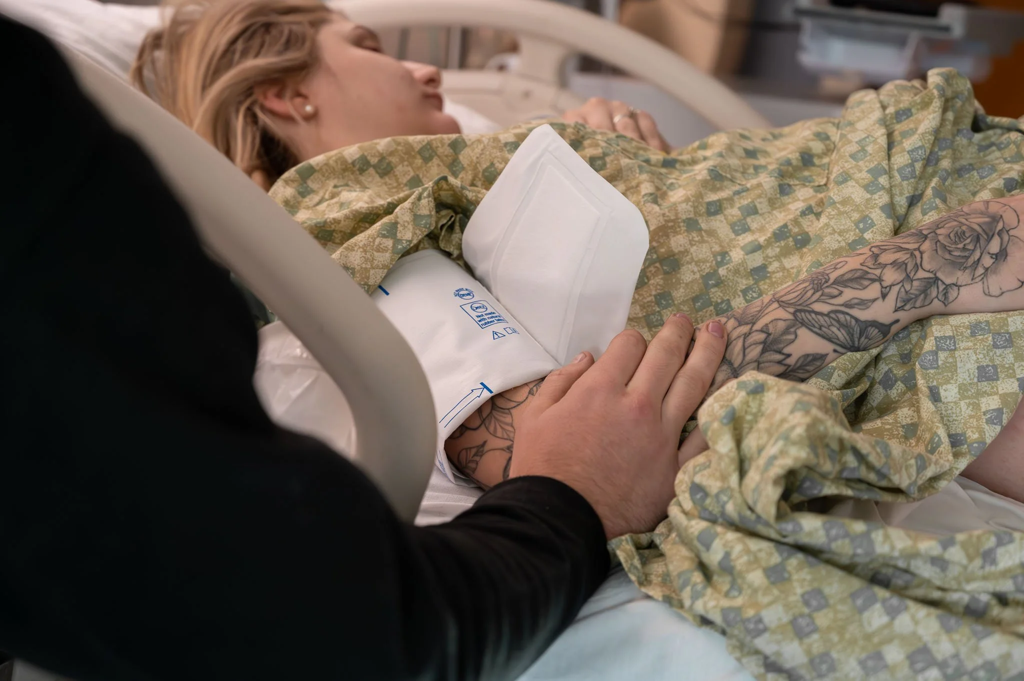 A woman lying in a hospital bed with a medical gown and tattoos on her arm, a healthcare professional is holding her hand.