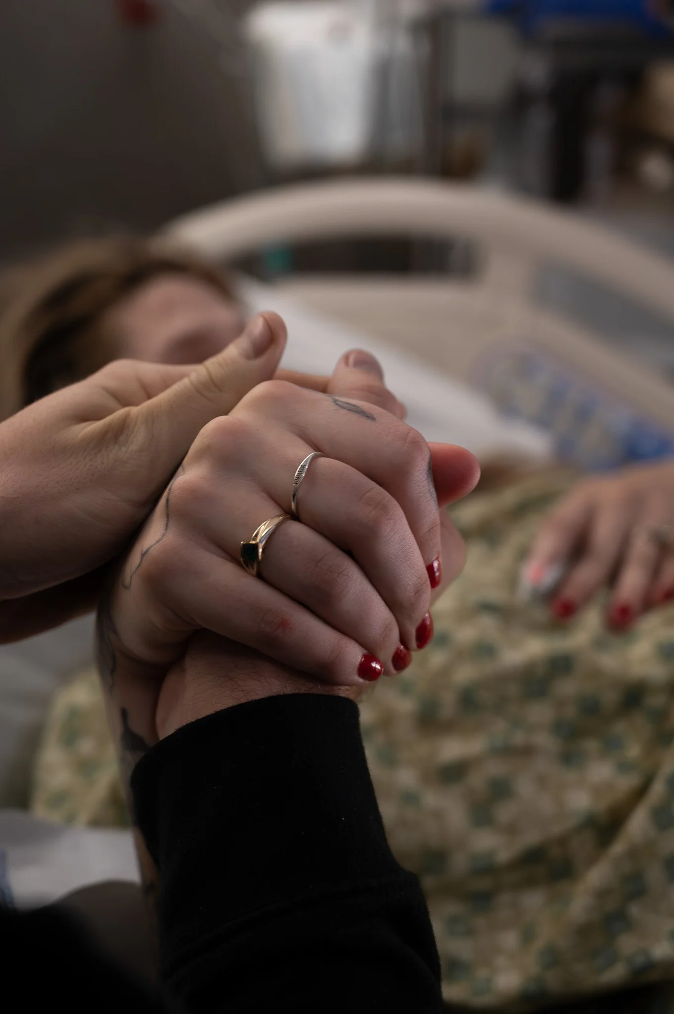 Two people holding hands, with one wearing rings, in a hospital room with a patient lying in a bed in the background.