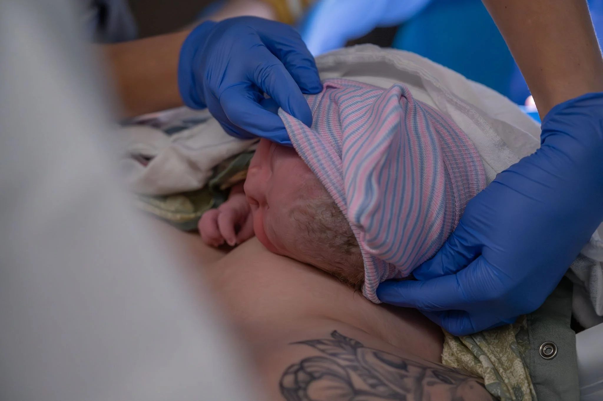 A newborn baby wearing a striped hospital cap being gently held and examined by medical staff with blue gloves, lying on a person's chest.