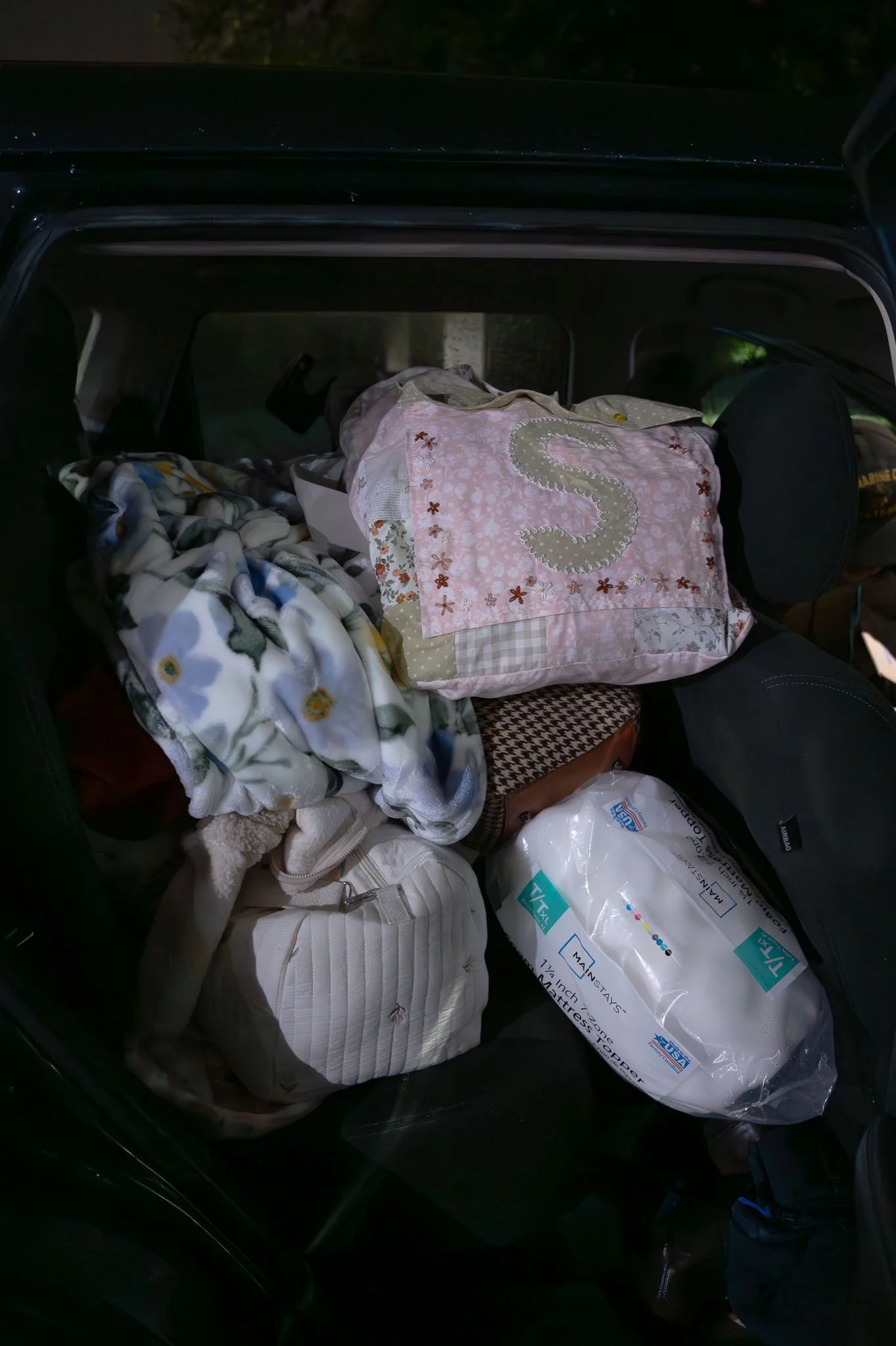 Back of a car packed with blankets, bags, a pillow, and a mattress protector.