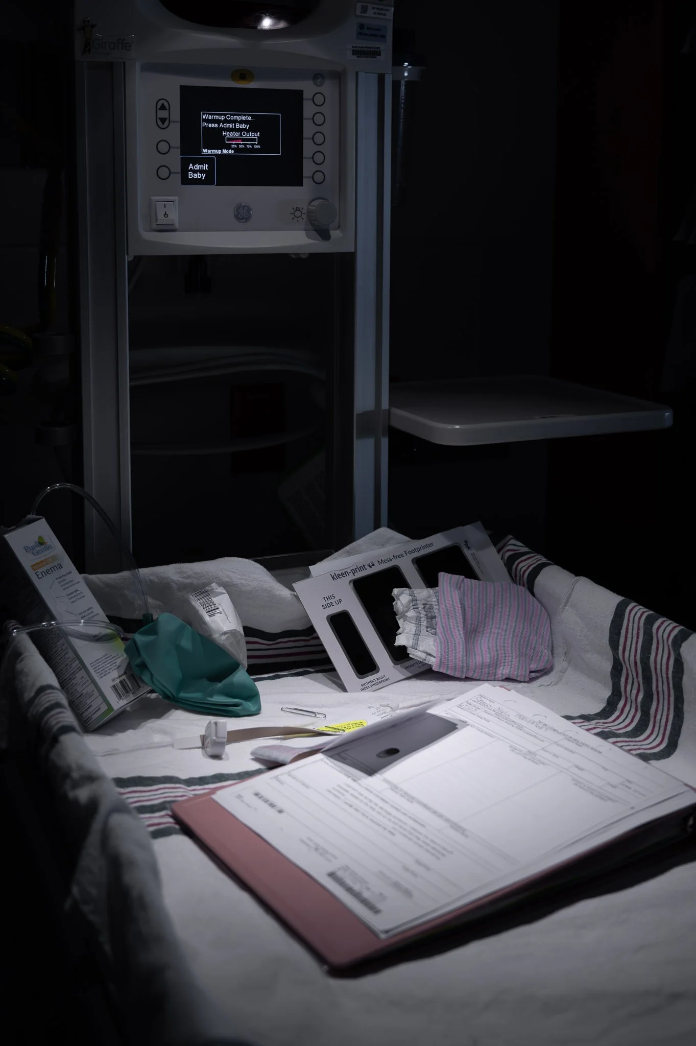 A hospital room with a NICU incubator, medical supplies, a medical chart, and a foot pedal for a medical device in a dimly lit environment.