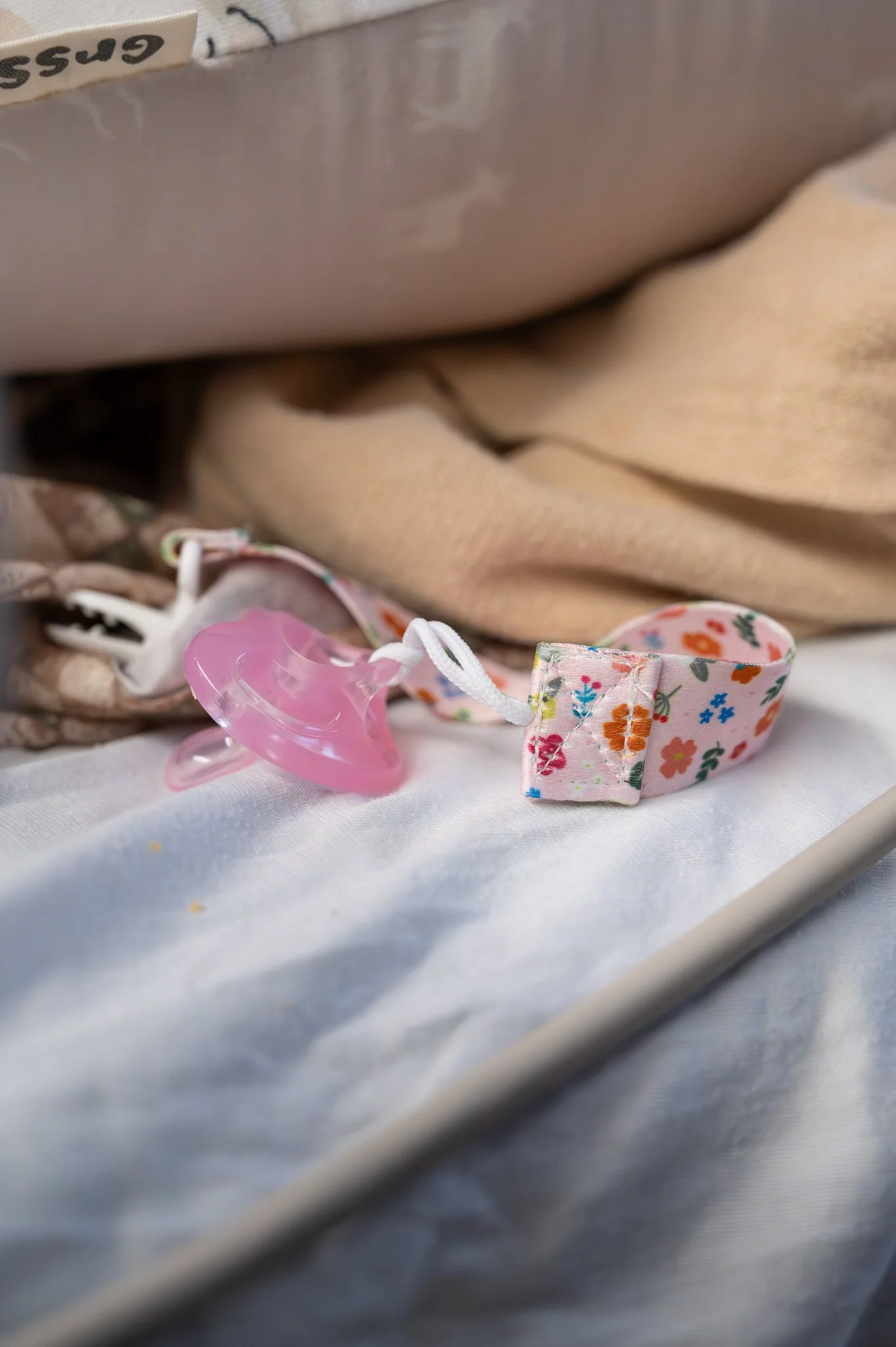 A pink pacifier with a floral fabric strap on a hospital bed with beige blankets.