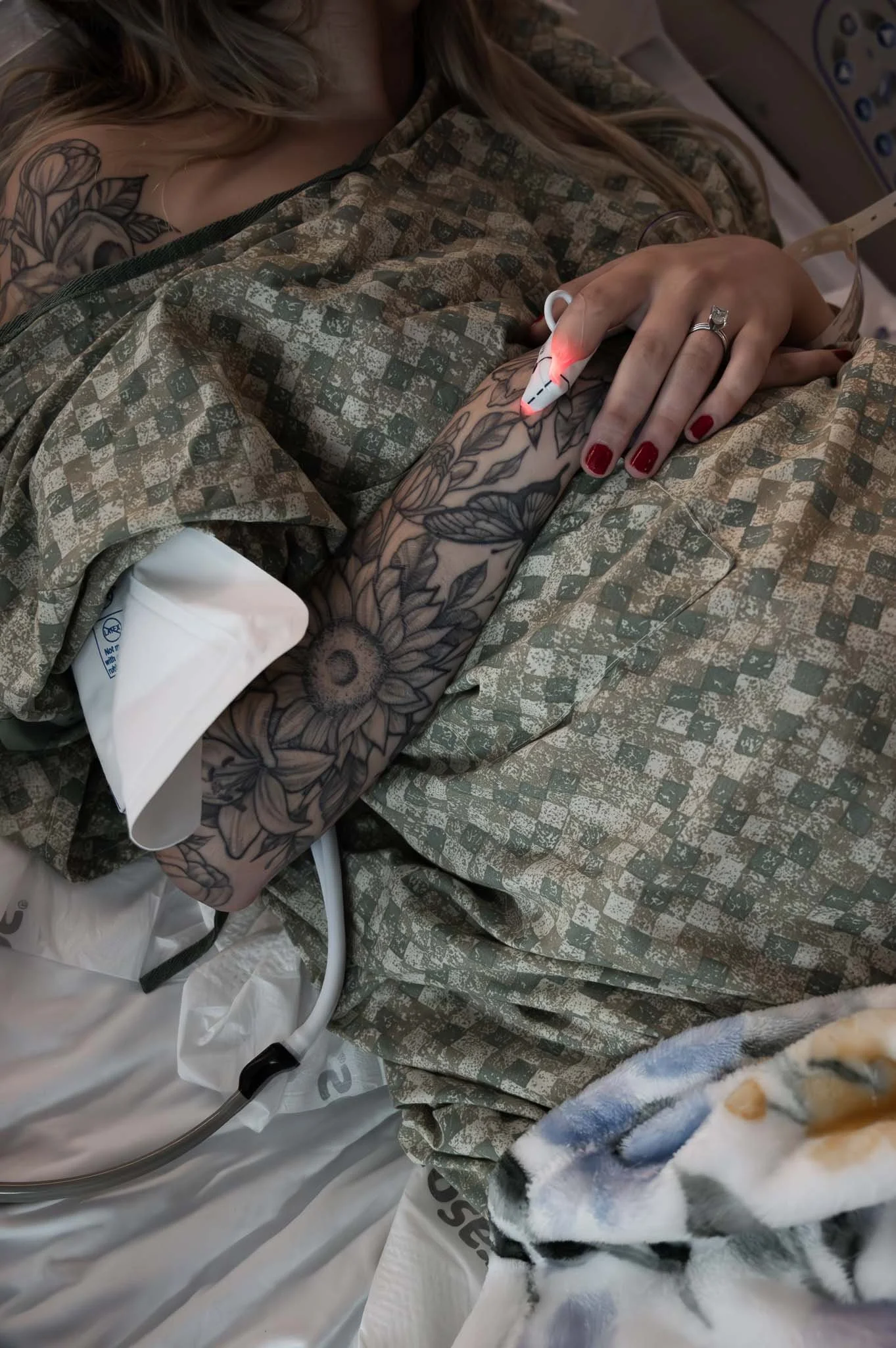 A woman in a hospital bed with tattoos on her arm, holding a pulse oximeter on her finger, showing her in a medical setting.