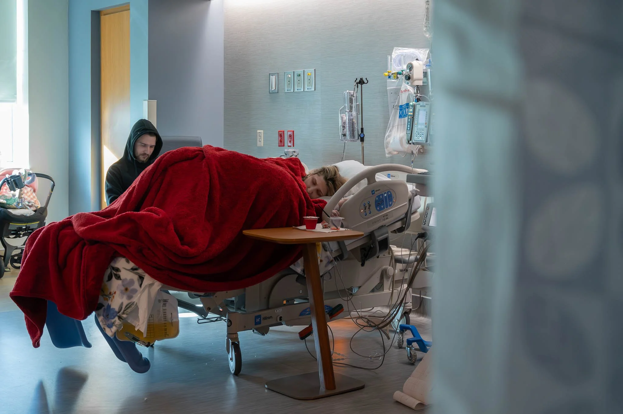 A hospital room with a patient lying in bed covered with a red blanket, a person sitting beside the bed, media equipment on the wall, and medical devices nearby.