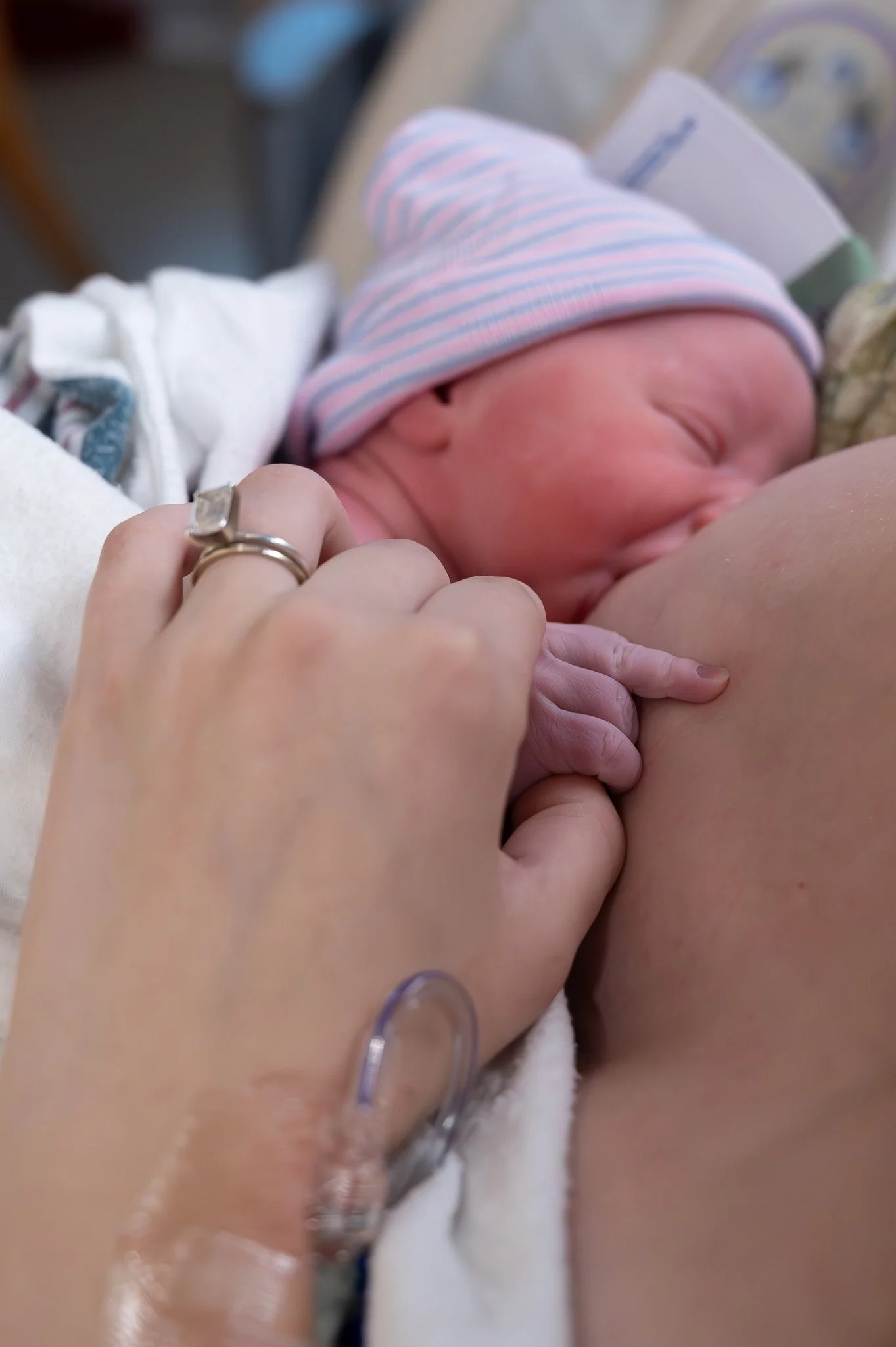 A newborn baby in a striped hat sucking on a mother's breast, with a hospital wristband and wedding ring visible.