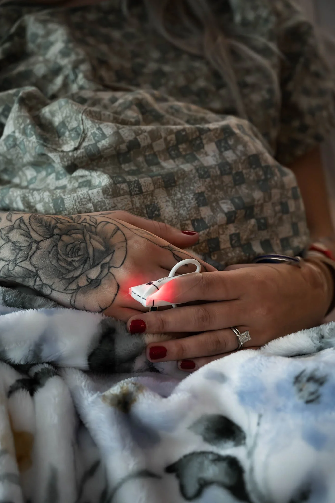 A hand with detailed rose tattoo holding a medical device emitting red light, resting on a plush blanket with black and white spots, and wearing a ring with a large stone.