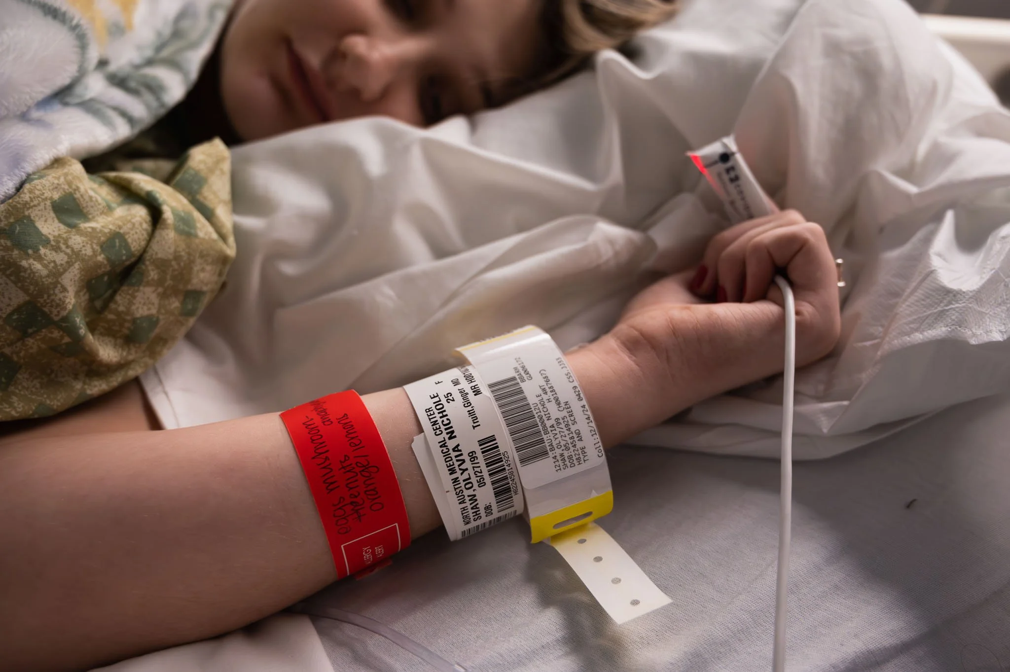 Close-up of a person lying in a hospital bed with an IV line in their hand, wearing a hospital wristband and a patterned blanket.