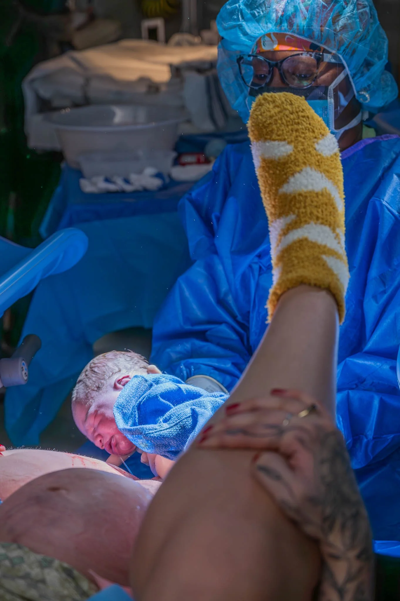 A healthcare professional wearing protective gear assists in childbirth, holding the newborn after delivery as the mother lies on the table.