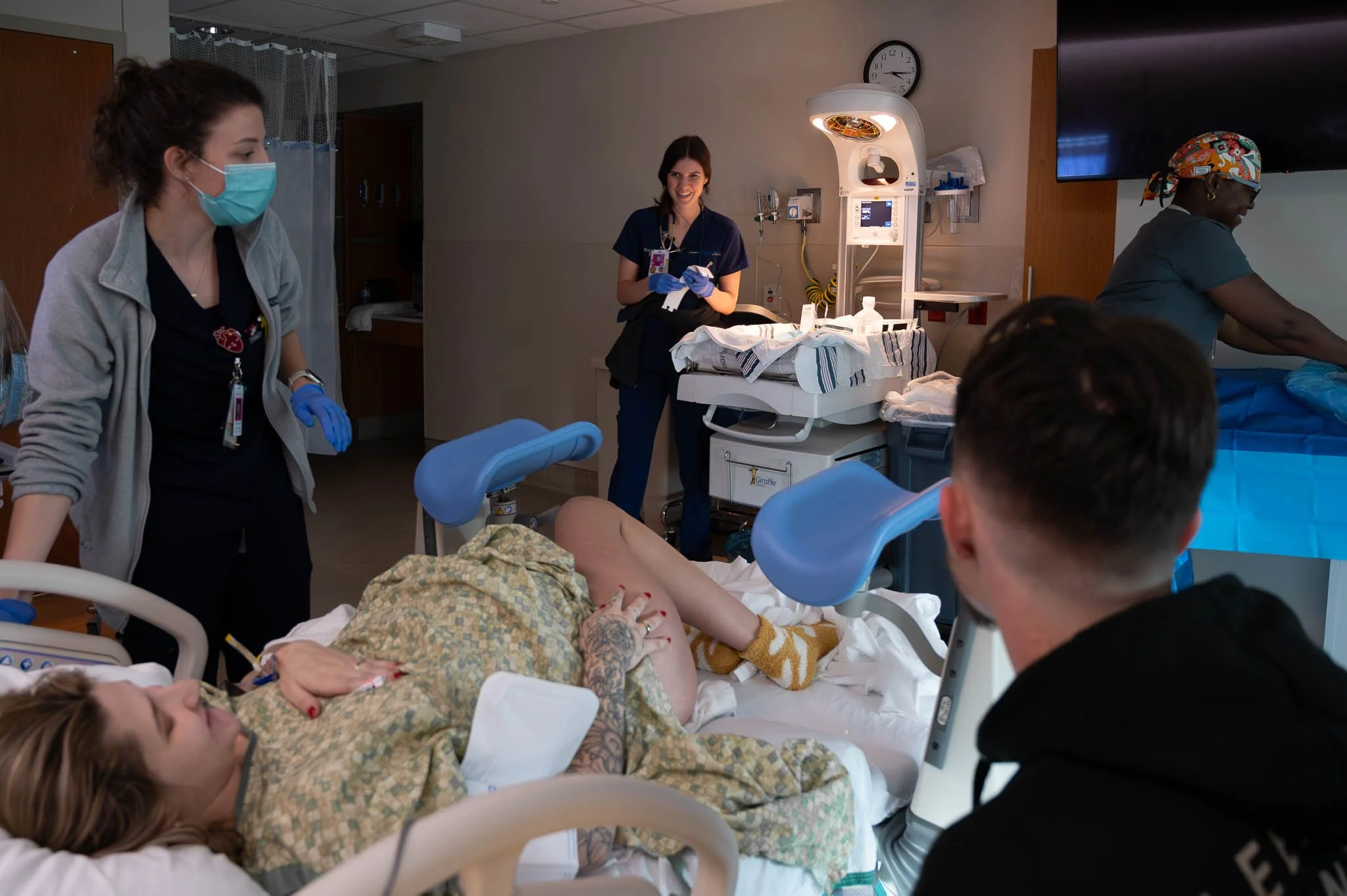 A woman lying in a hospital bed giving birth, with medical staff and visitors present in the hospital room.