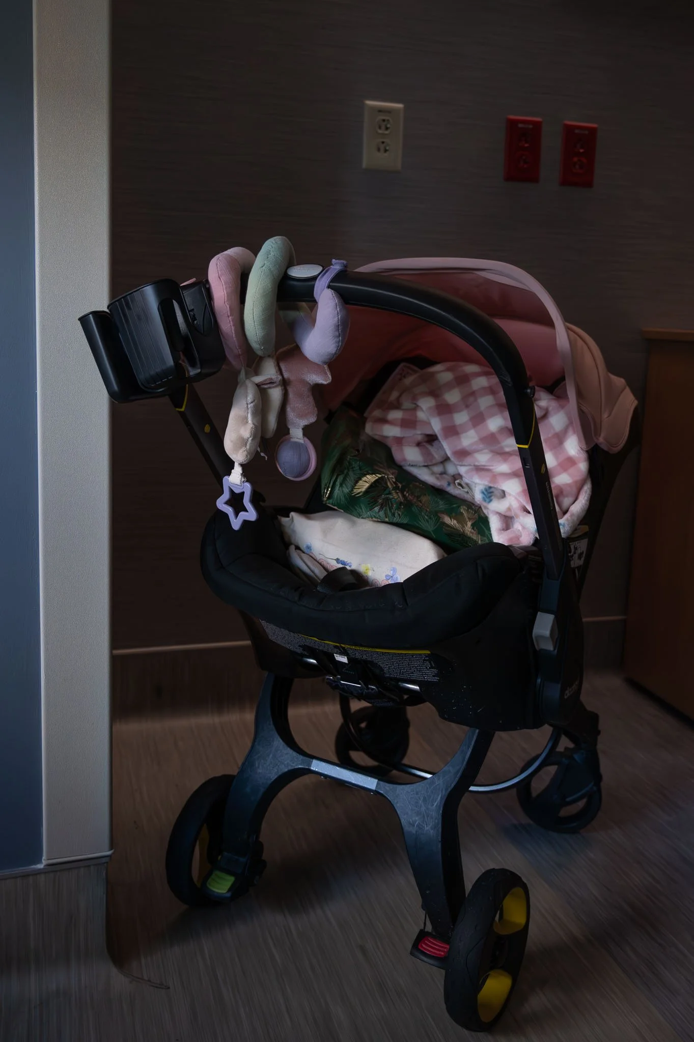 A stroller with stuffed animals hanging from the handle, containing blankets and other items, situated on a wooden floor next to a wall with electrical outlets.