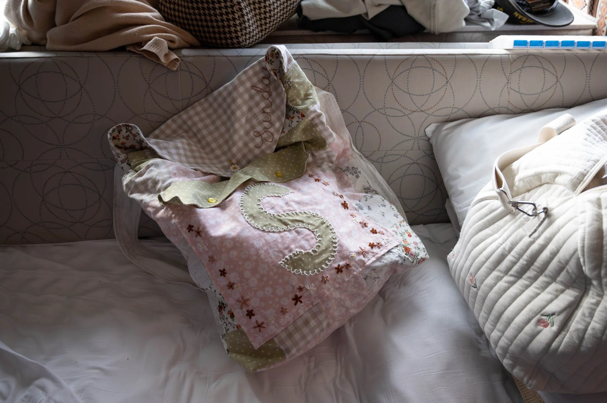 A bed with a pillow and a quilted bag on it, and a headboard with a patterned cover. There are various items on a shelf or surface at the head of the bed.