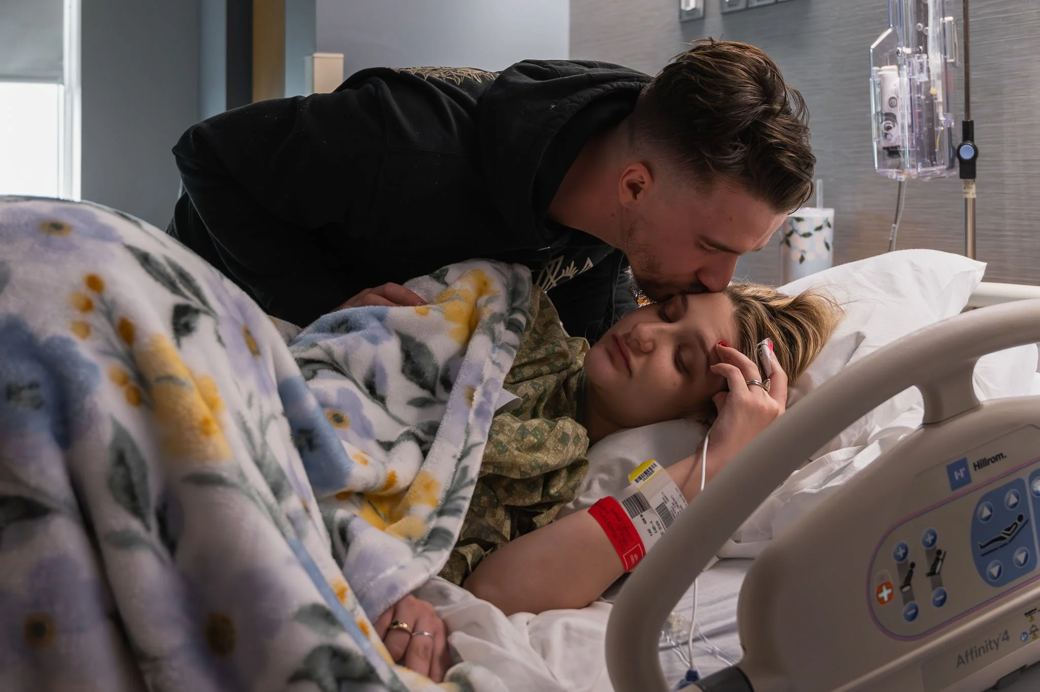 A man kisses a woman lying in a hospital bed, who appears to be recovering from an illness. The woman is resting with her eyes closed, wearing a hospital gown and a blanket, and has a hospital wristband and IV in her arm.