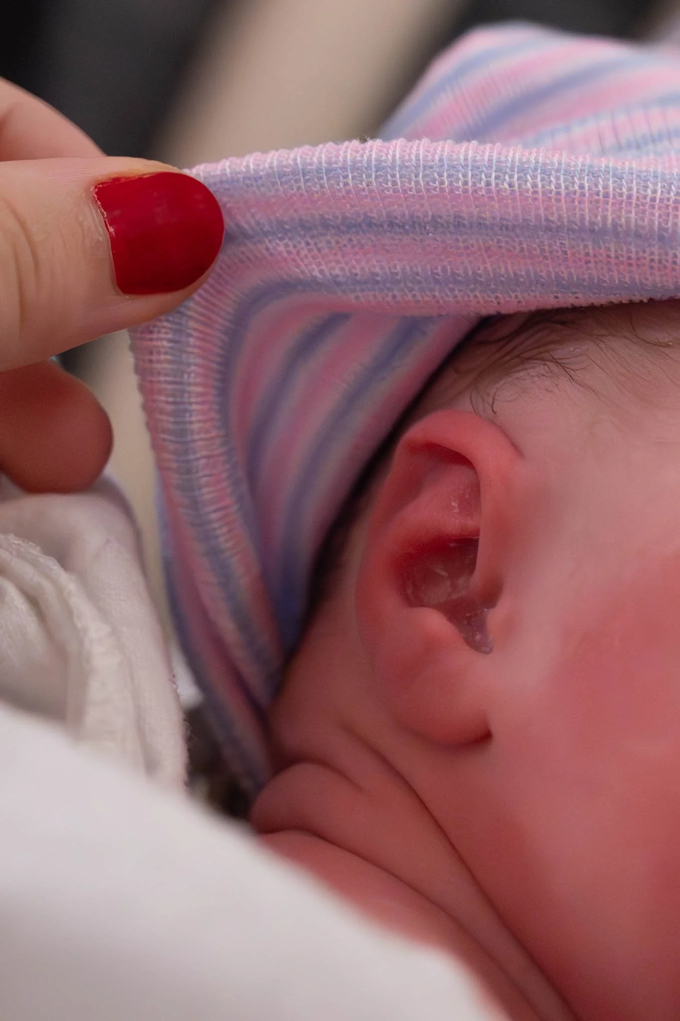 Close-up of a person's hand with red nail polish holding a baby's head, showing the baby's ear and part of a colorful striped hat.