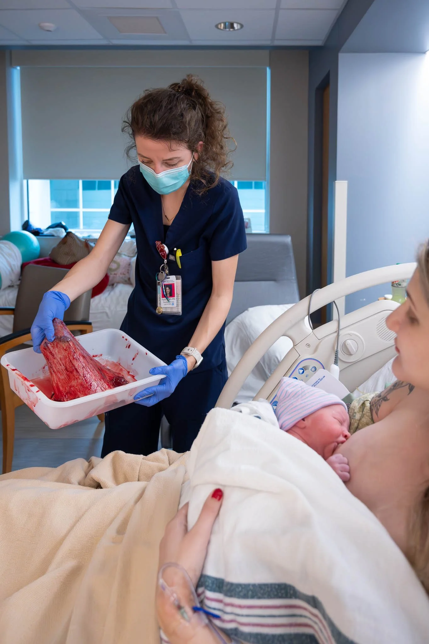 A nurse wearing a face mask and gloves holds a tray of umbilical cord blood. A mother is lying in a hospital bed with a newborn baby breastfeeding, in a hospital room with large windows.