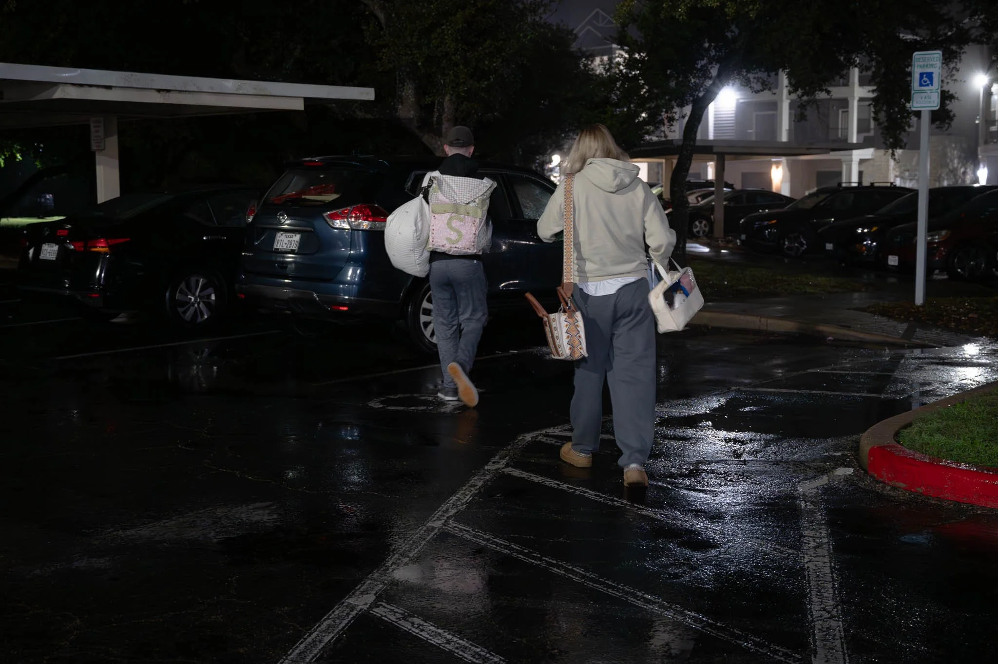 Two people walking through a wet parking lot at night, carrying bags, with parked cars and a residential building in the background.