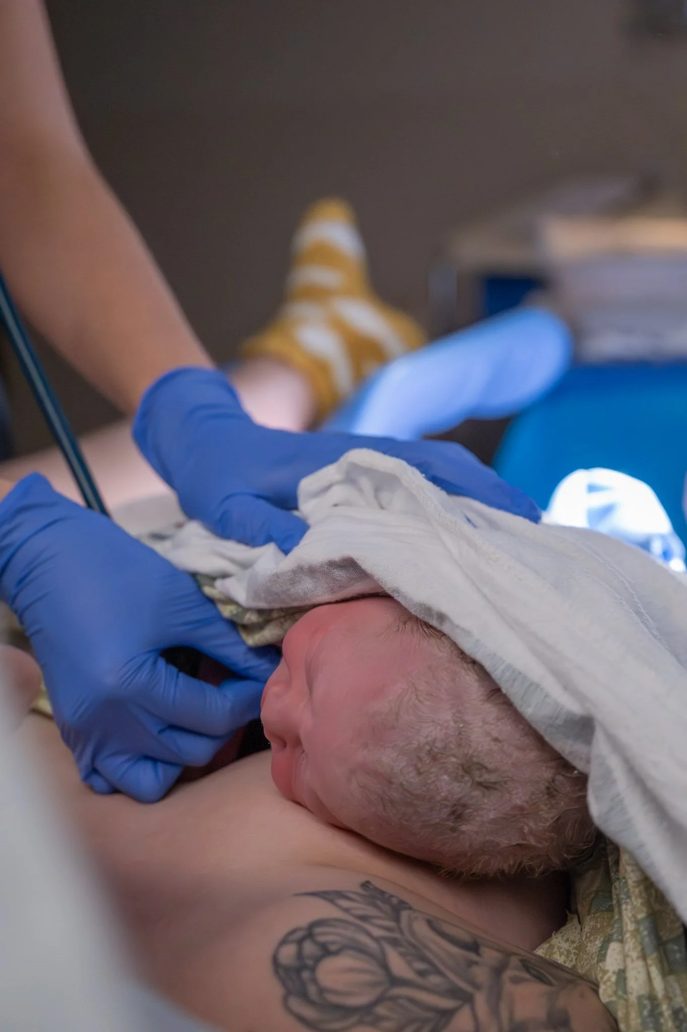 Newborn baby being delivered by a healthcare professional wearing blue gloves, with a towel over the baby's head, in a hospital setting.