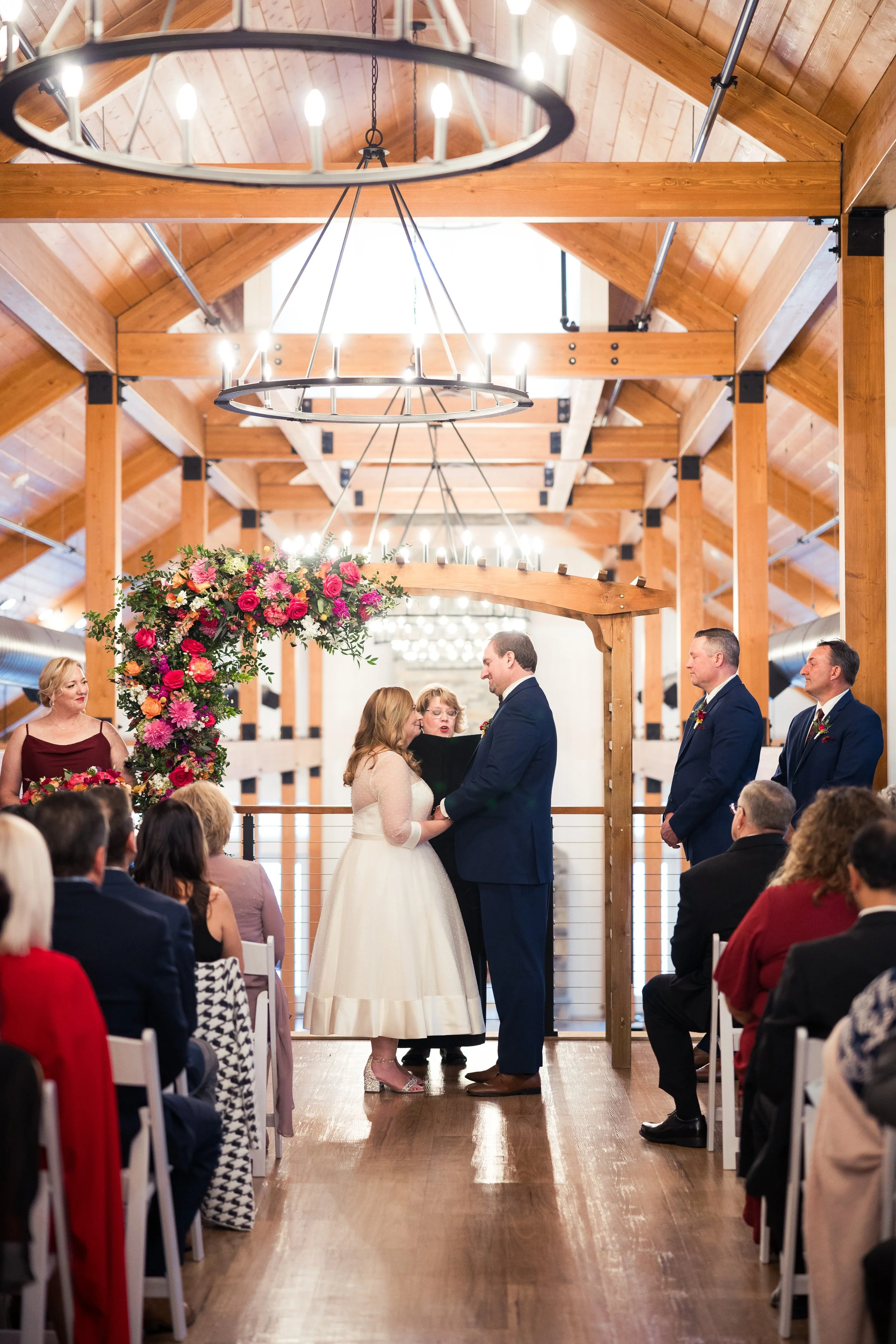 A couple stands before an officiant in a wooden barn, exchanging vows during a wedding ceremony. The bride wears a white dress and the groom wears a navy suit. Guests are seated on either side, watching the ceremony, with a floral arrangement of pink