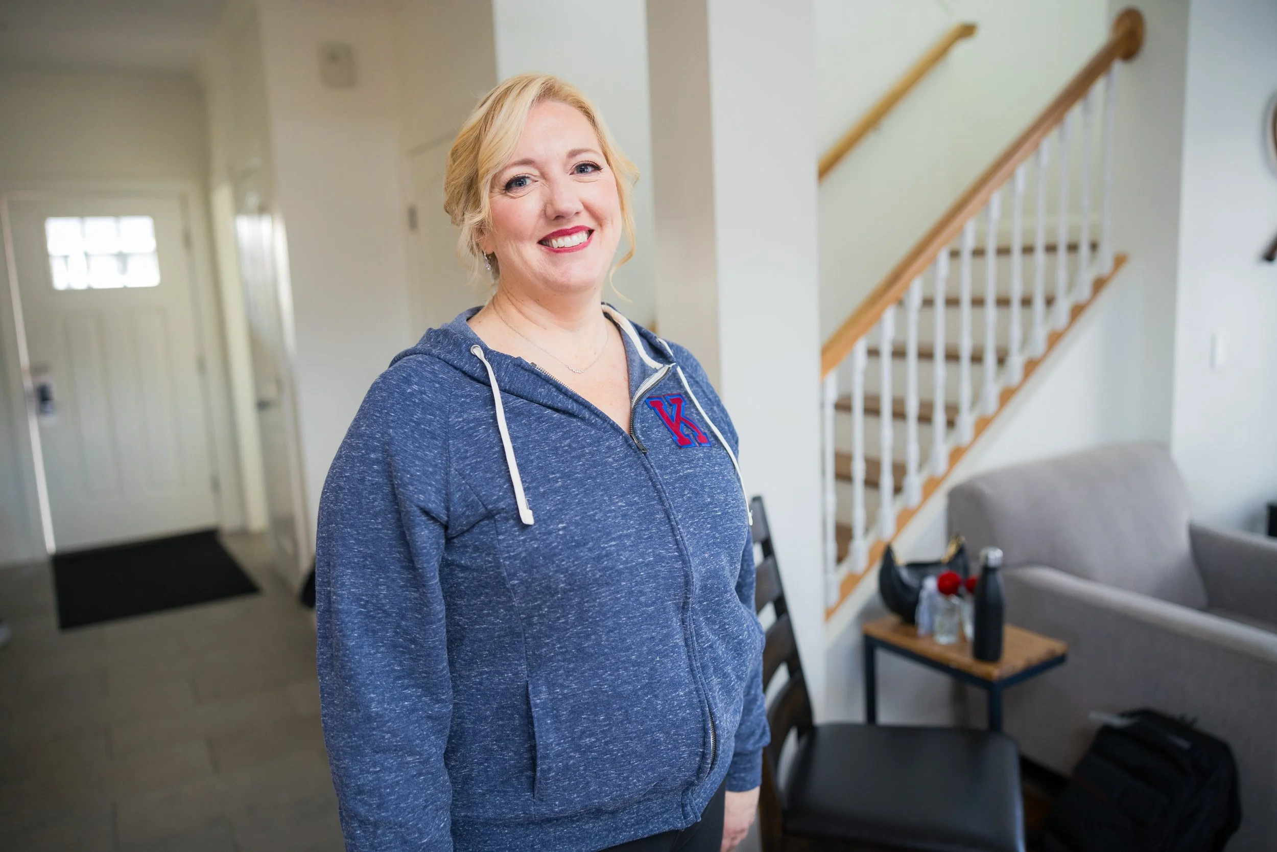 A smiling woman with blonde hair wearing a blue zip-up hoodie with a red and blue letter 'K' on it. She is indoors near a staircase with wooden handrails, in a room with white walls and furniture.