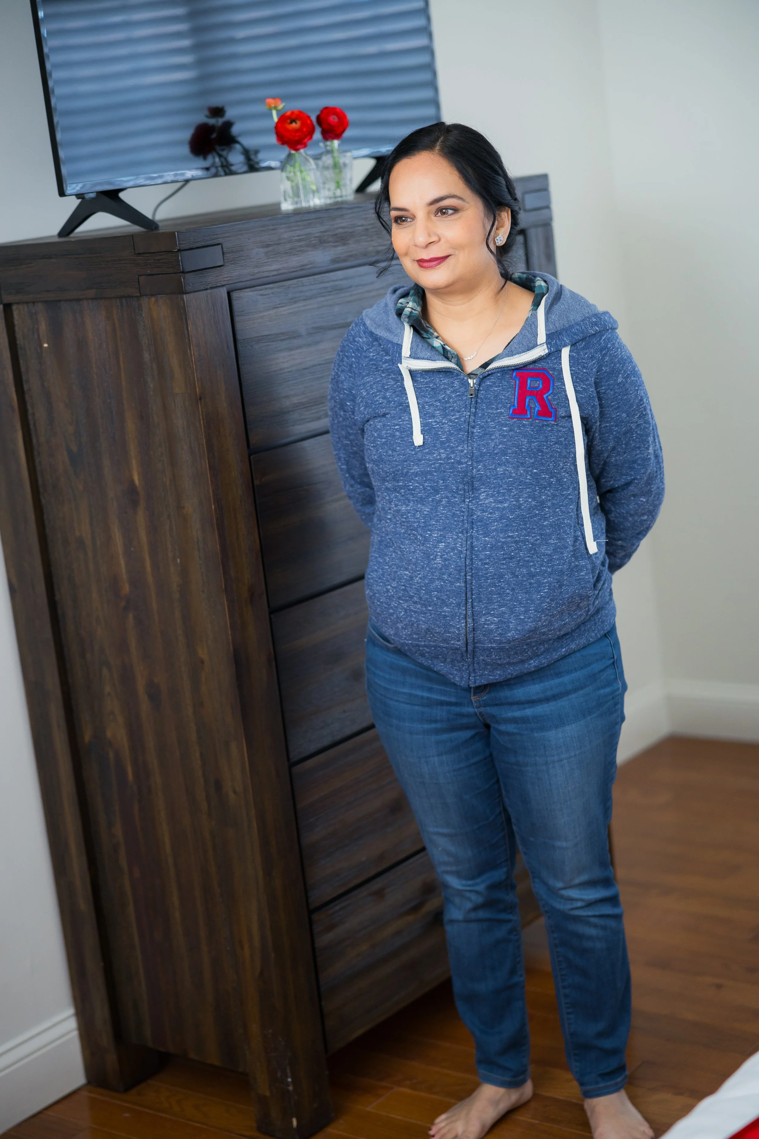 A woman standing barefoot by a wooden cabinet with a TV on top, smiling slightly, wearing a blue zip-up hoodie with a red and blue letter "R" on it, and blue jeans, with a vase of red and orange flowers on the TV stand in the background.