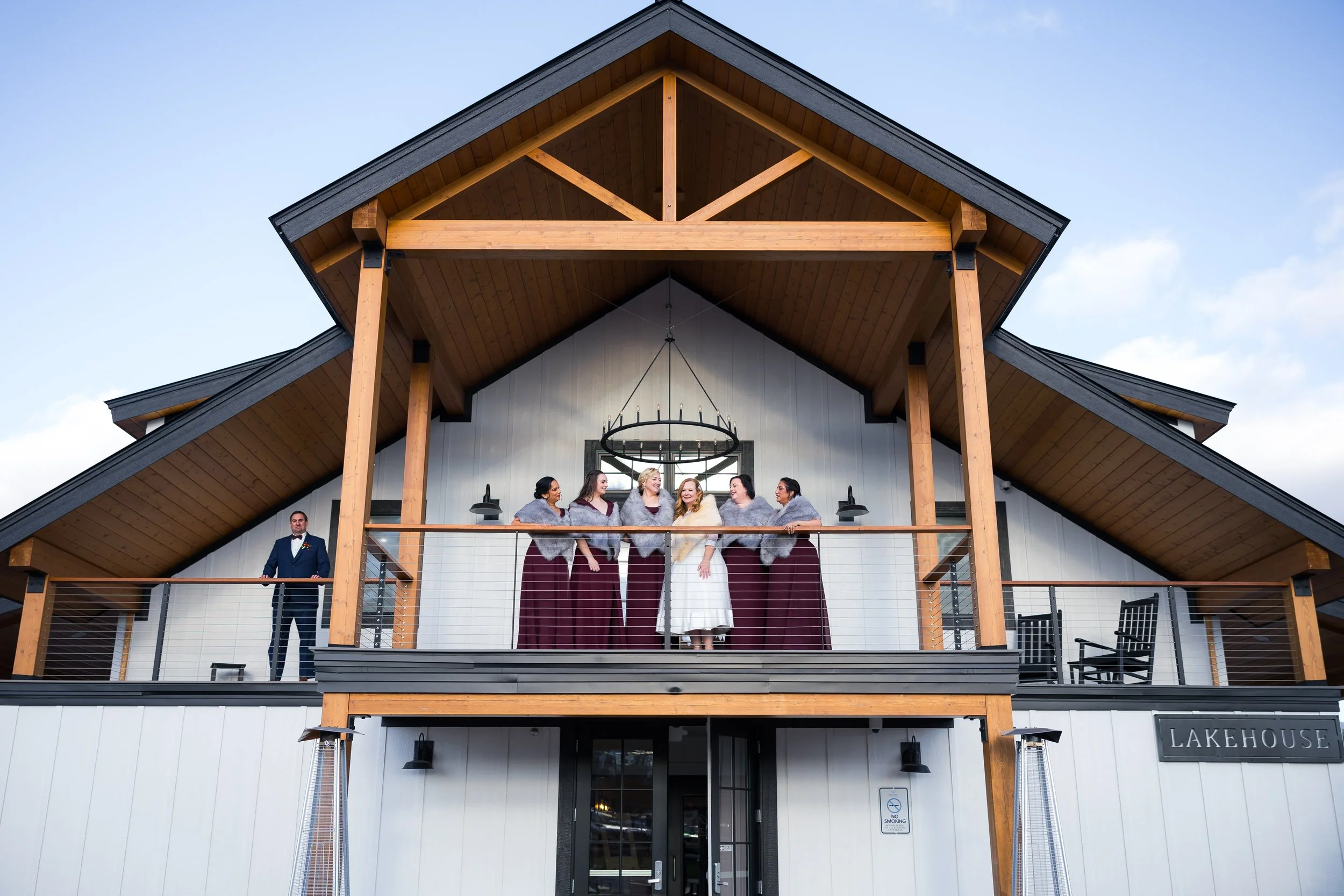 Wedding group photo on a balcony of a modern lakehouse building with a wooden roof design. Bride in white gown and bridesmaids in maroon dresses with gray shawls, groomsman in a suit, all smiling and talking.